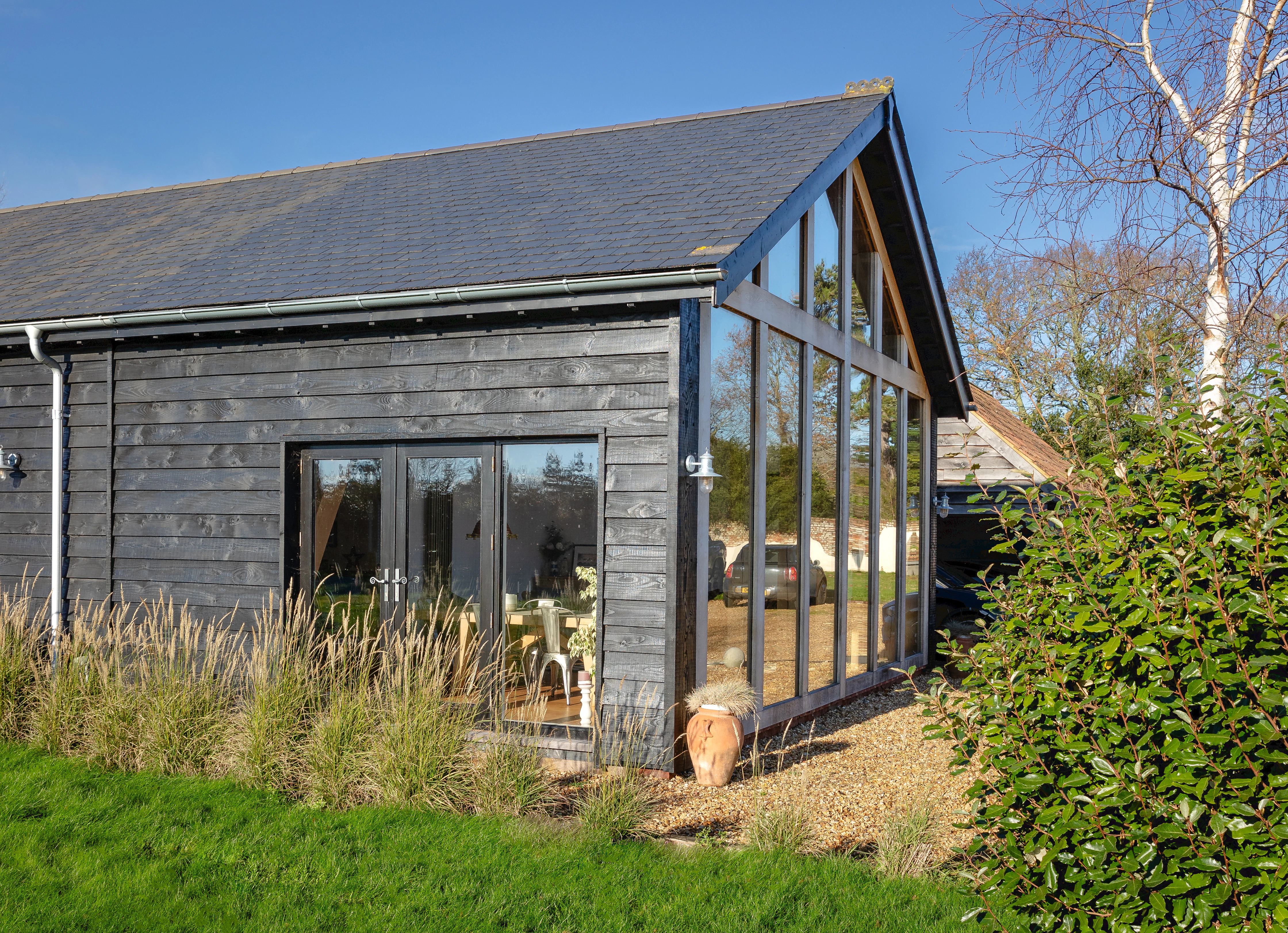 The side of an oak framed extension with a glazed gable and external black cladding showing some of the garden with a green lawn and tall grasses