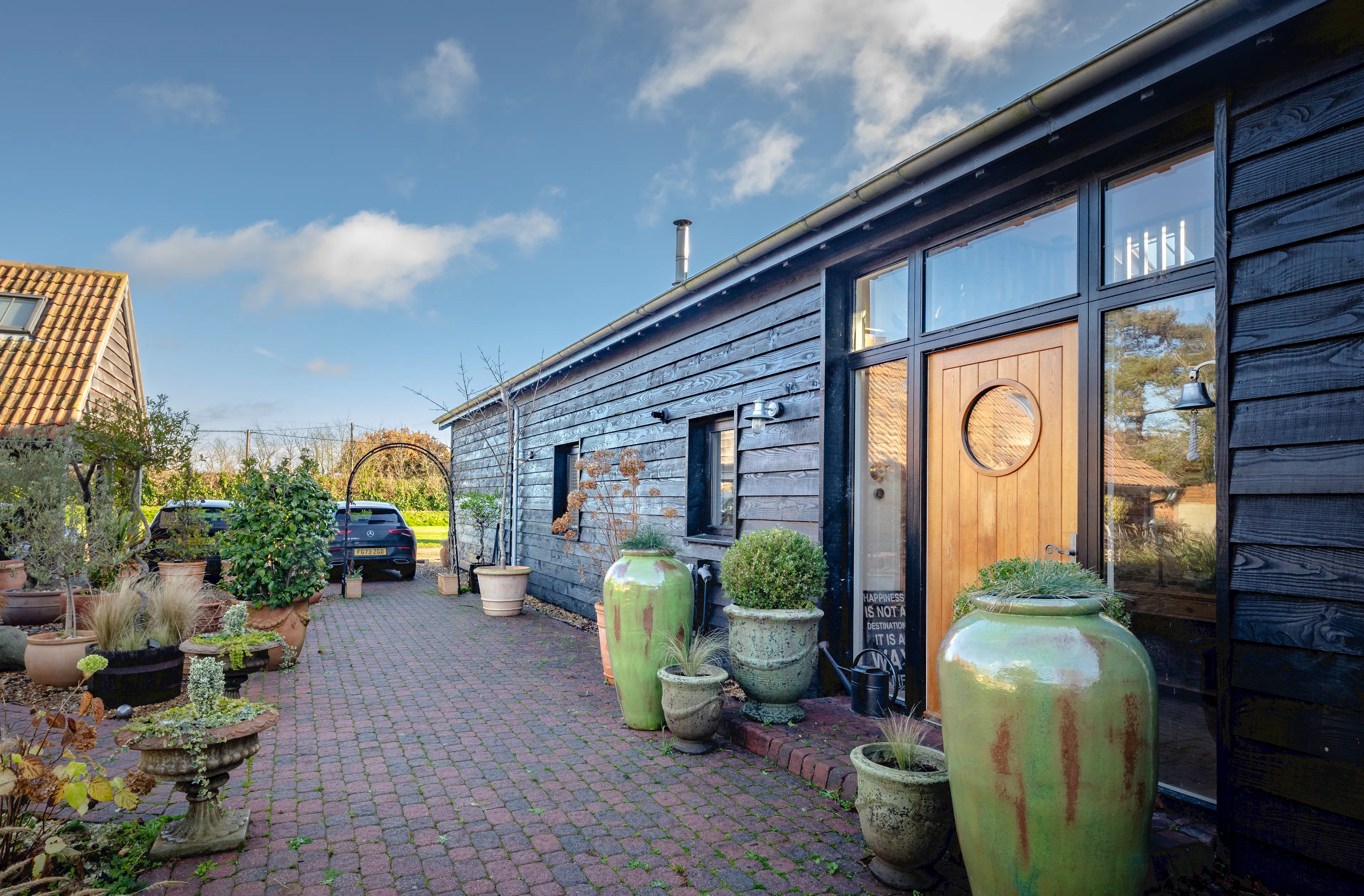 The side of an oak framed extension with a glazed gable and external black cladding, showing a paved walkway, front door and large plant pots
