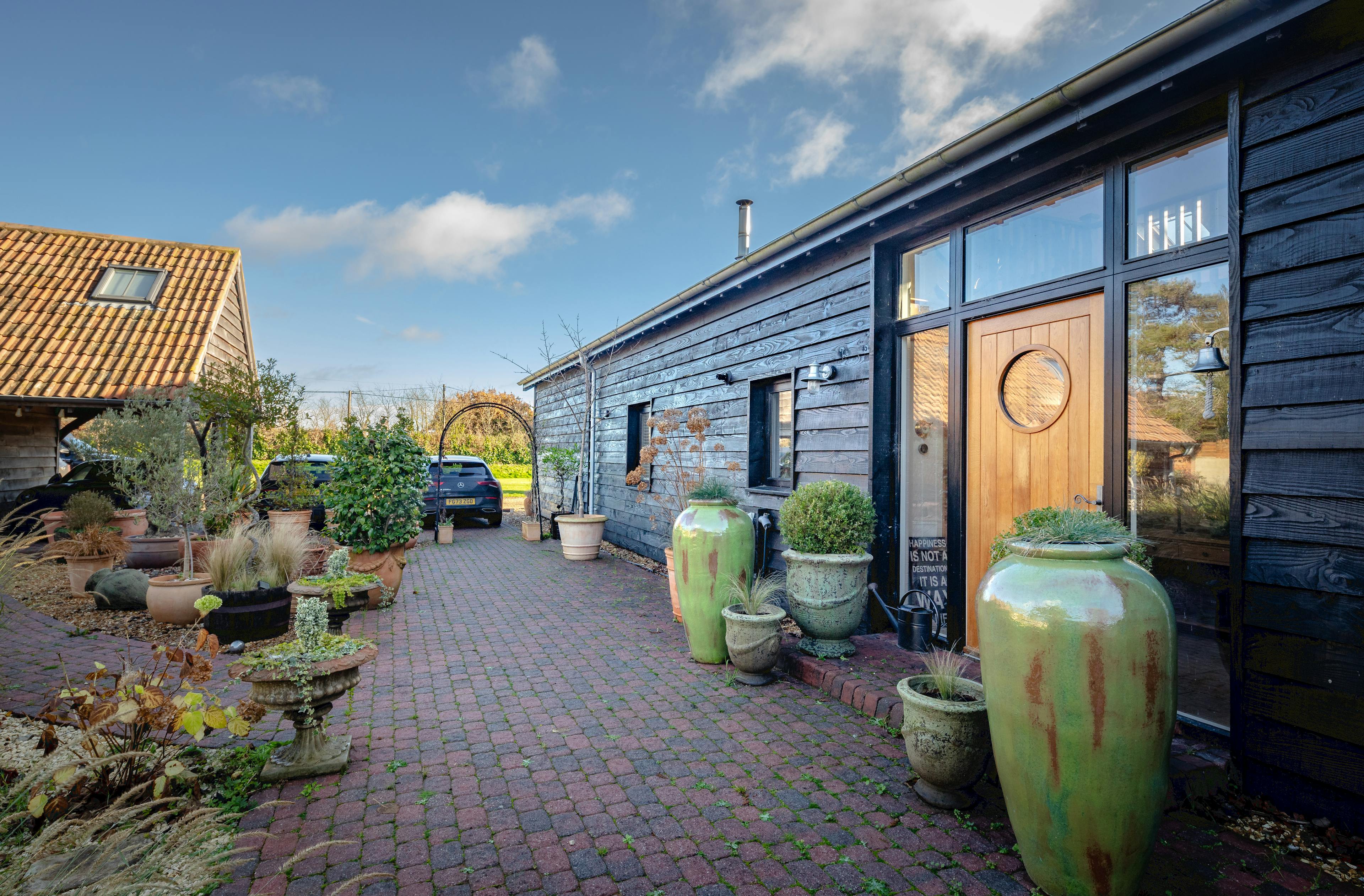 The side of an oak framed extension with a glazed gable and external black cladding, showing a paved walkway, front door and large plant pots