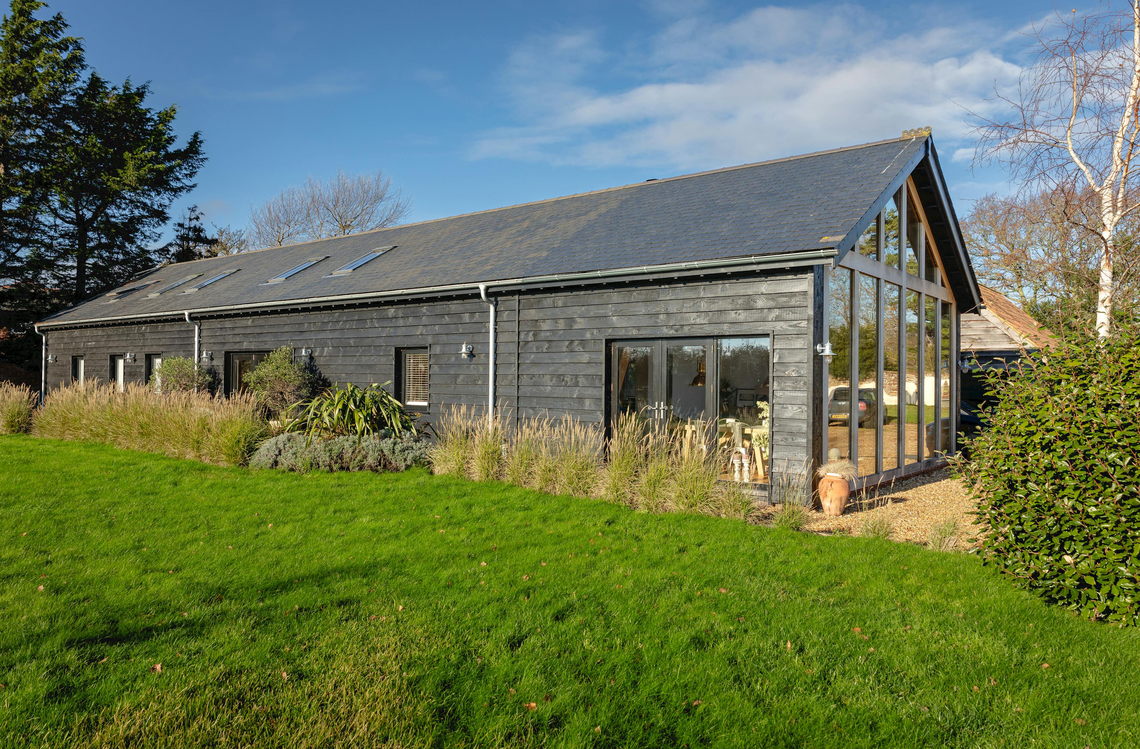 The side of an oak framed extension with a glazed gable and external black cladding showing some of the garden with a green lawn and tall grasses
