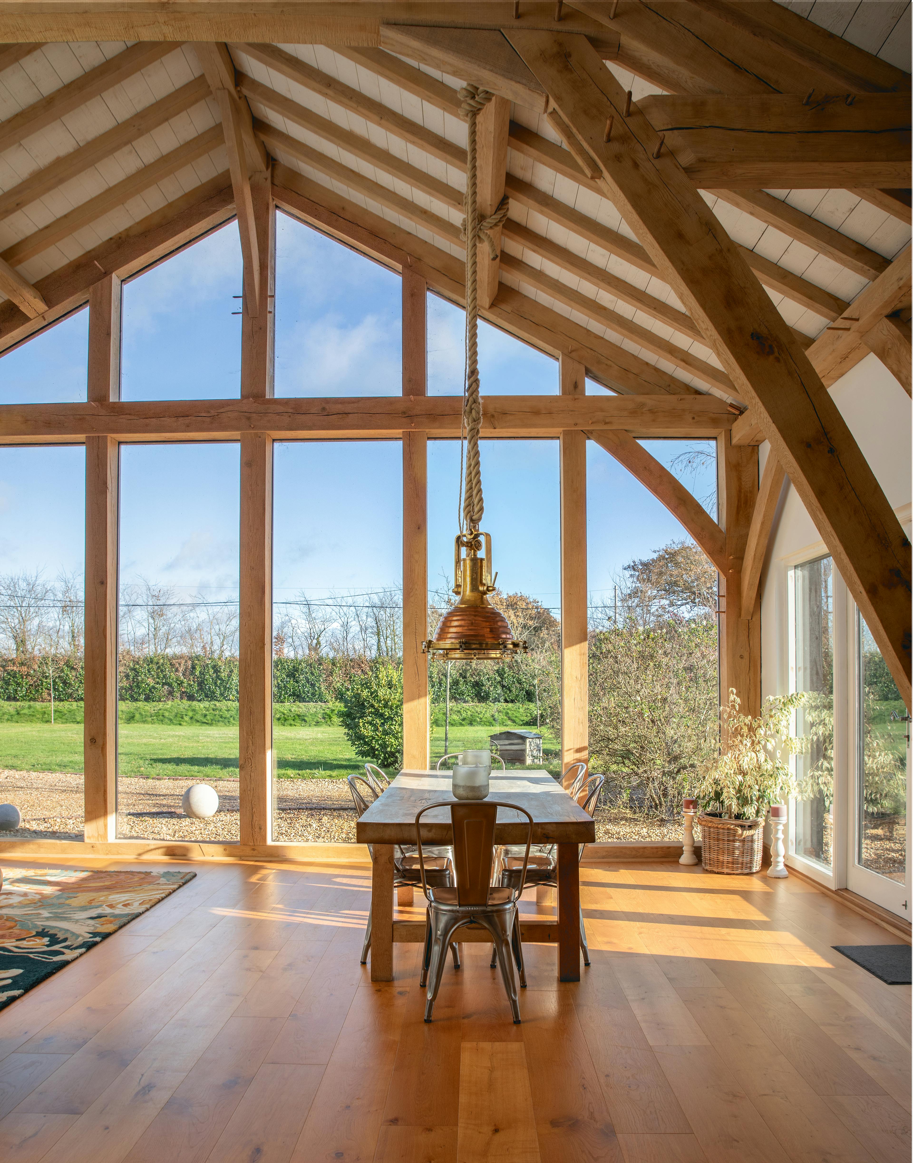 Sun beams shine through the windows on a wooden table and chairs and sideboard in an oak framed extension with a glazed gable