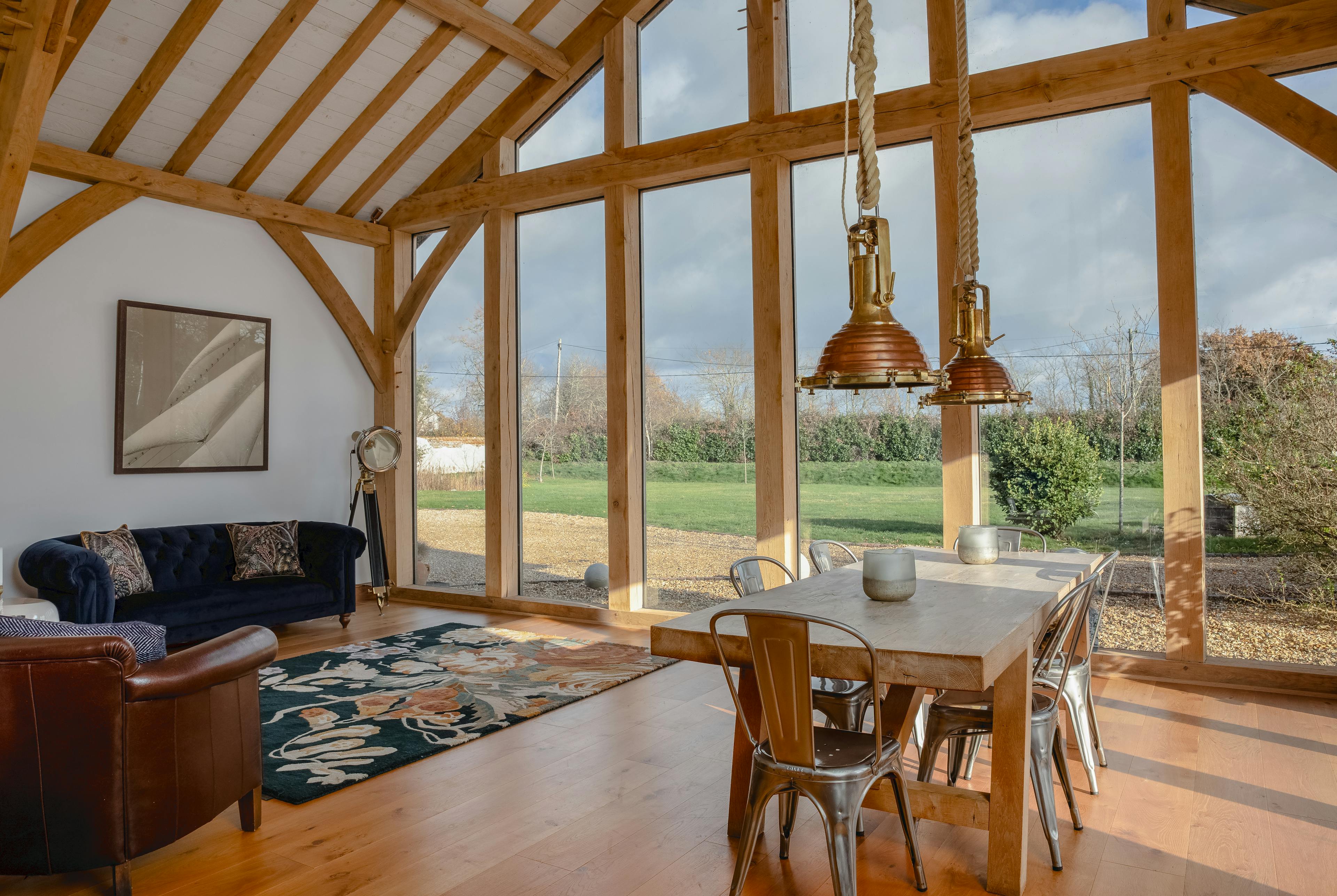 A wooden dining table and chairs, sofa and armchair in an oak framed extension with a glazed gable