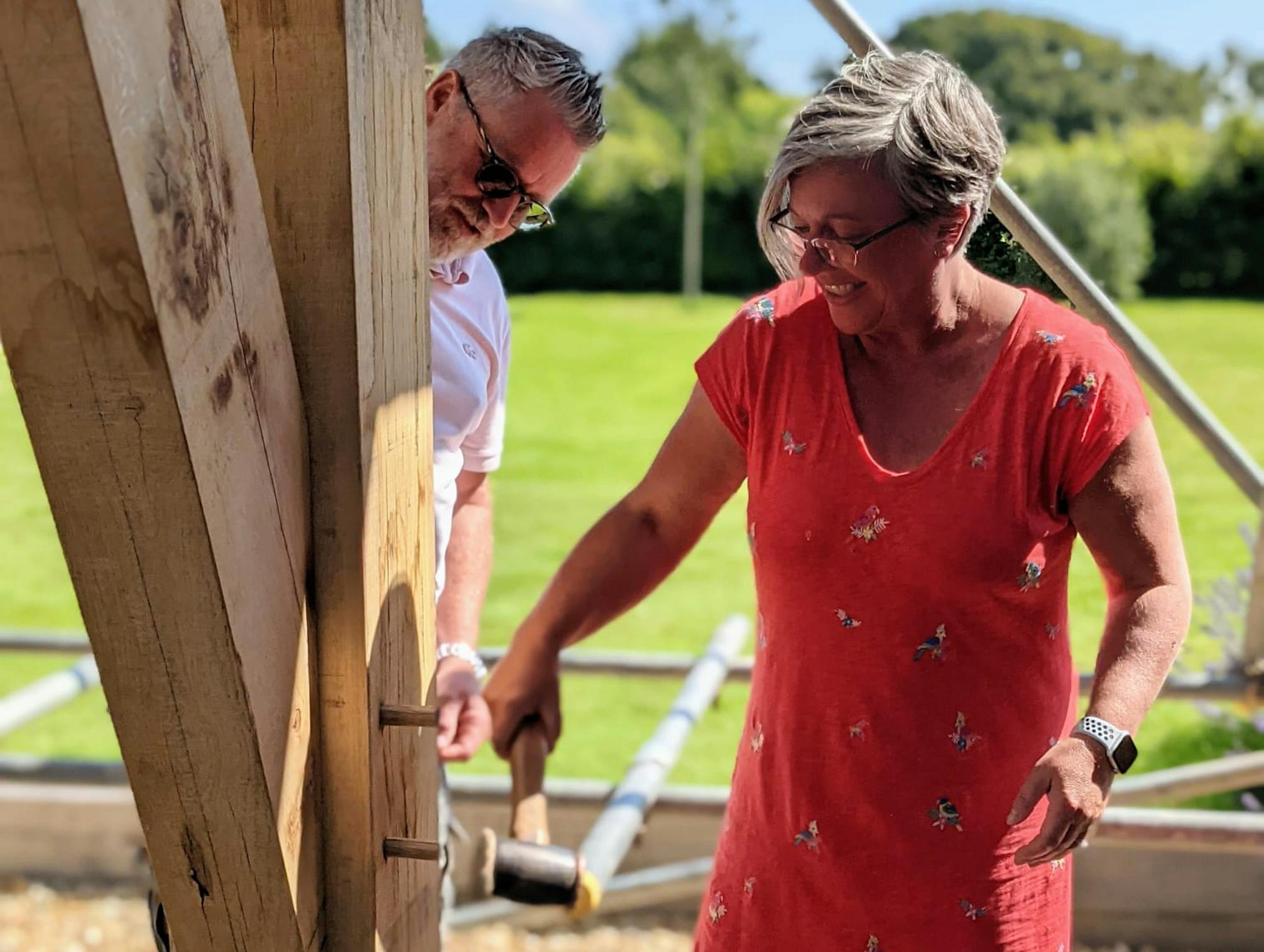 A woman in a red dress hammers a wooden peg into an oak frame