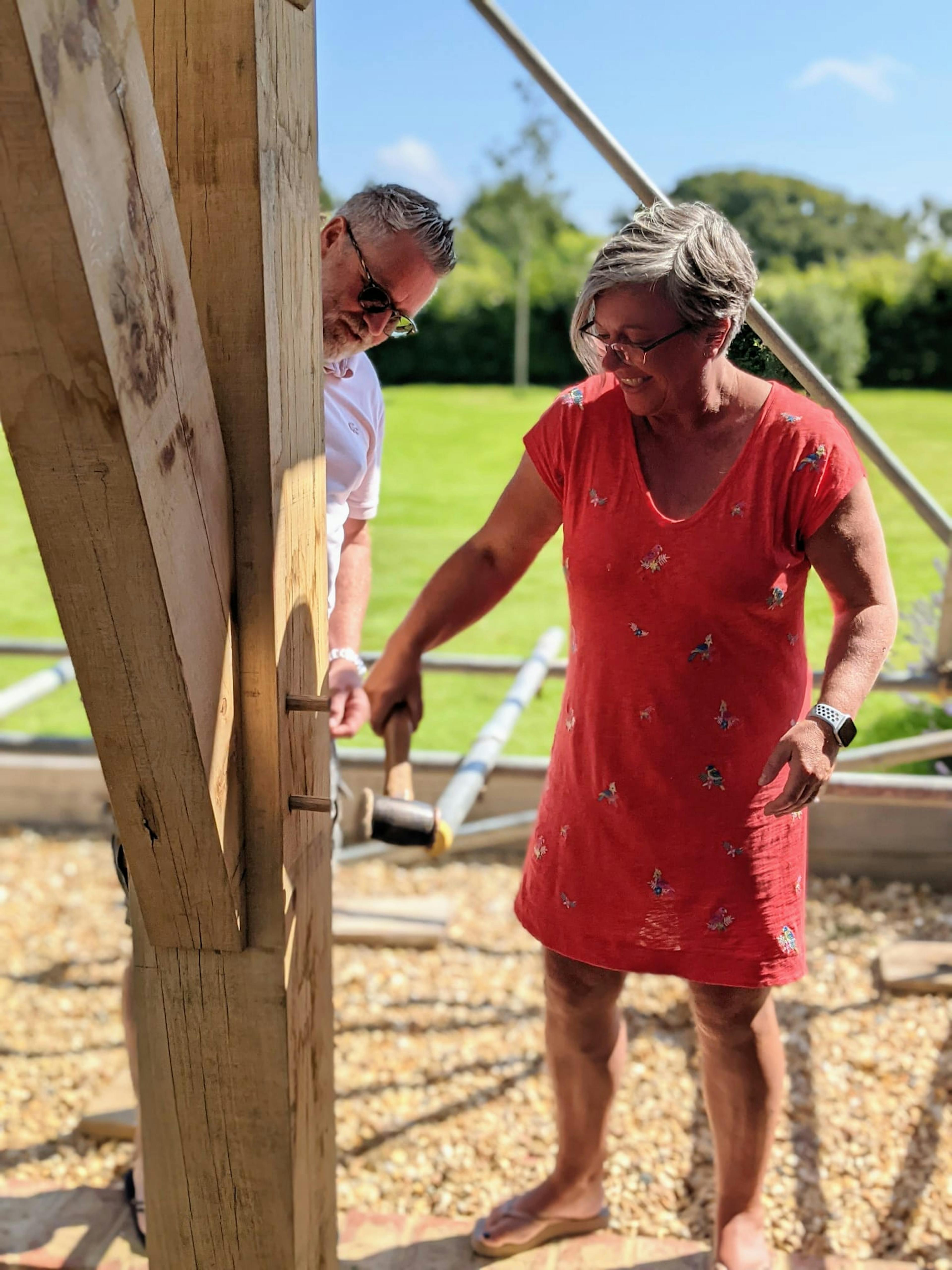 A woman in a red dress hammers a wooden peg into an oak frame
