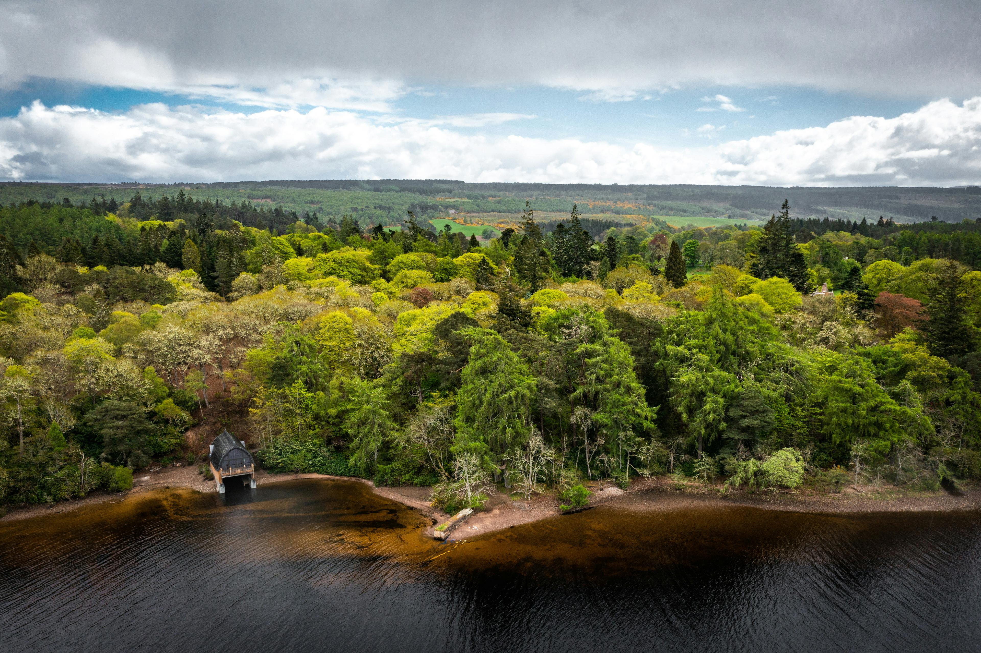 A view of a timber framed boathouse on loch ness with a curved roof with surrounding forests and Scottish hills