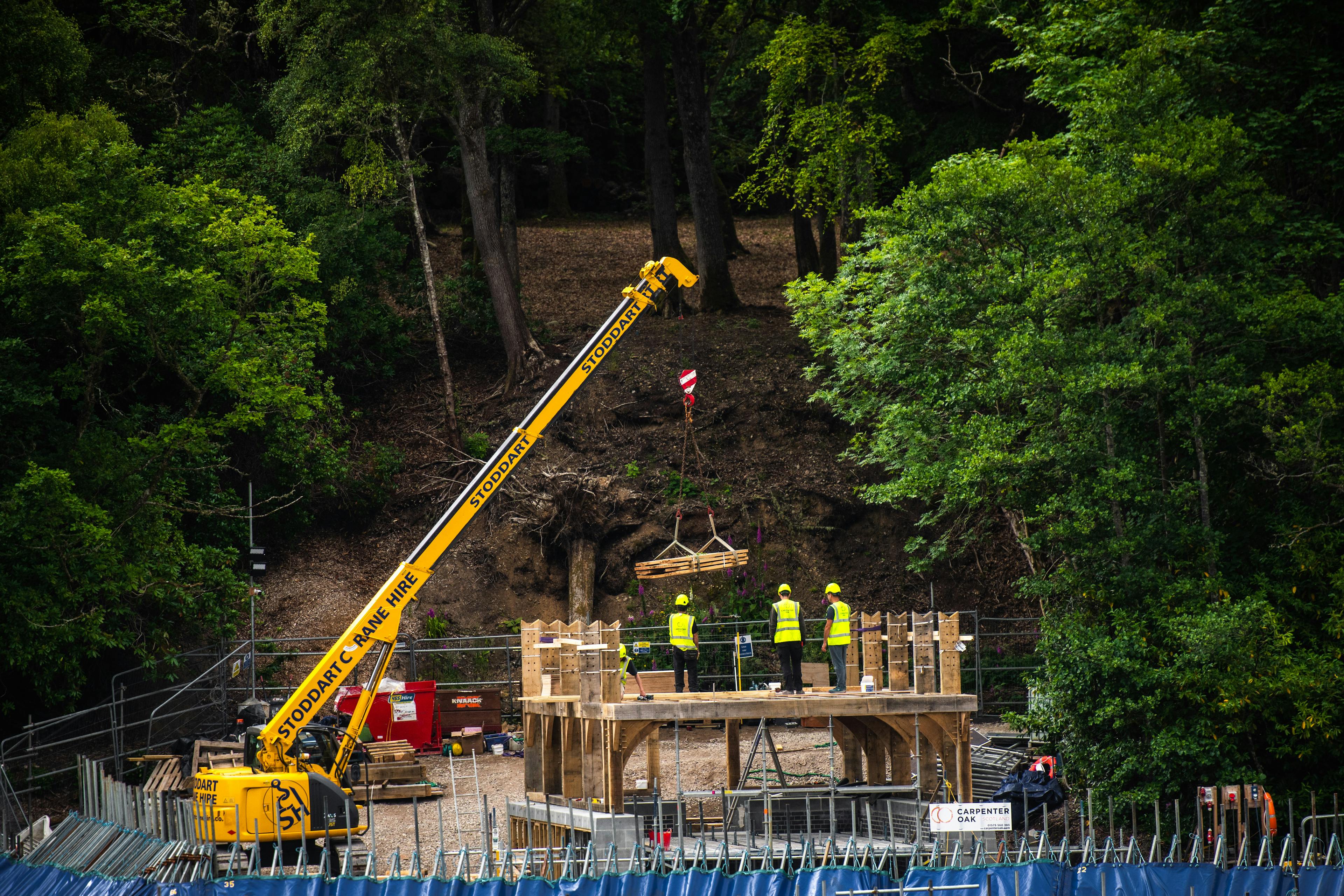 A yellow crane lifts into place the oak frame of a timber boathouse on loch ness with a curved roof during construction