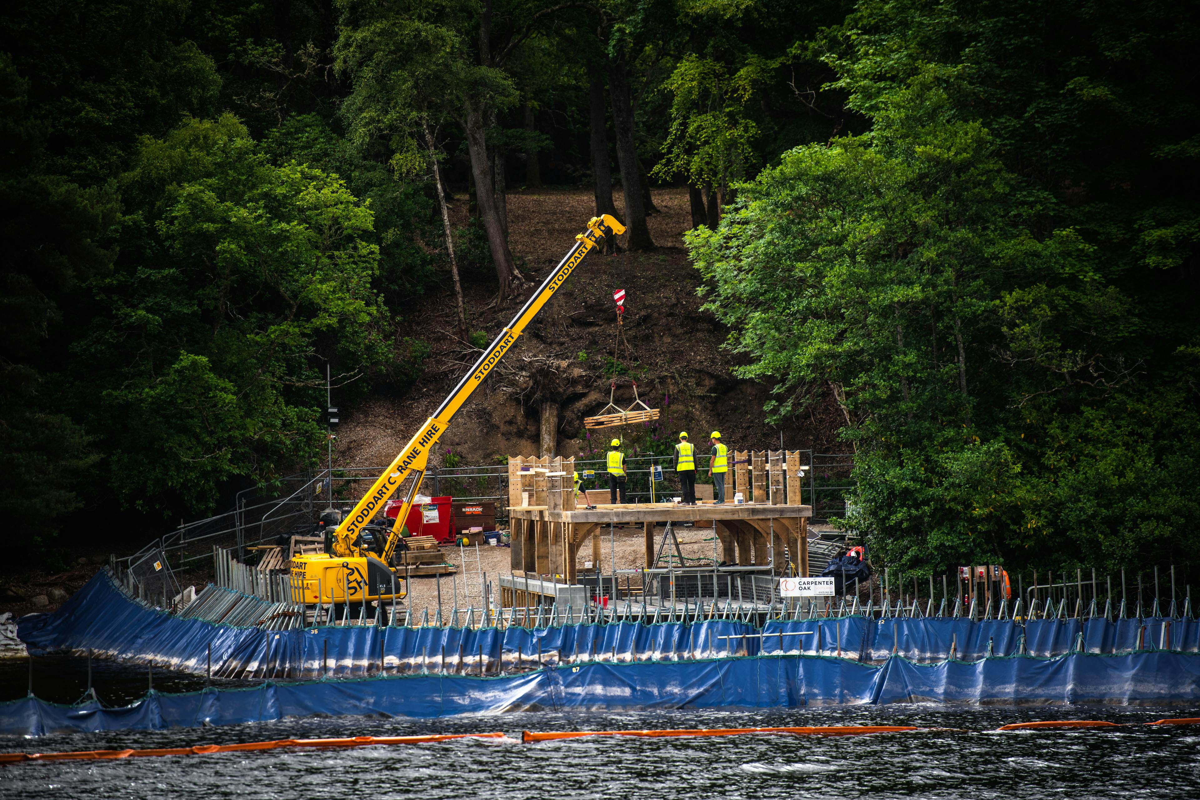A yellow crane lifts into place the oak frame of a timber boathouse on loch ness with a curved roof during construction