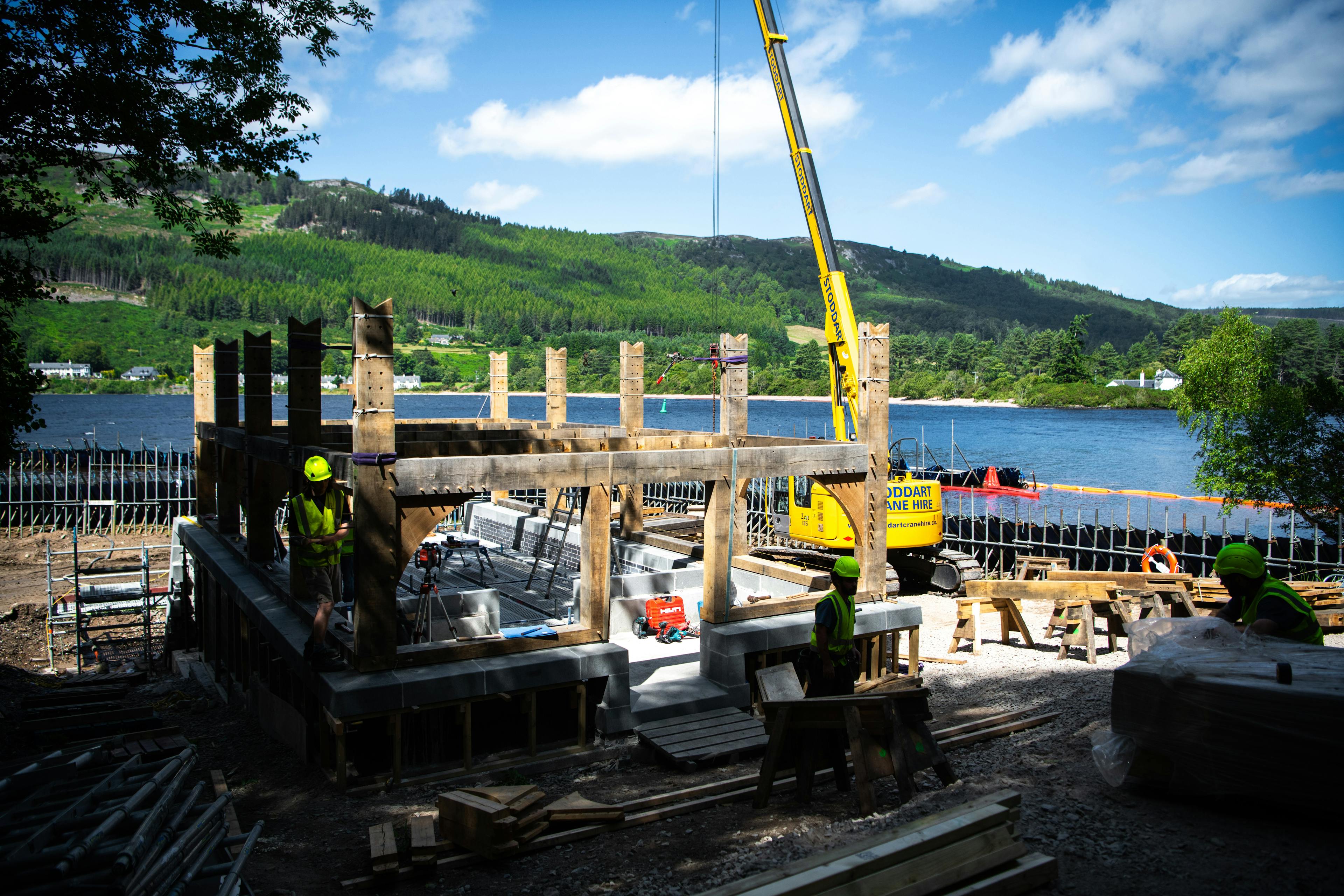 A view of a lake and surrounding green trees and hills with a lake side construction site for a timber framed boathouse in the foreground
