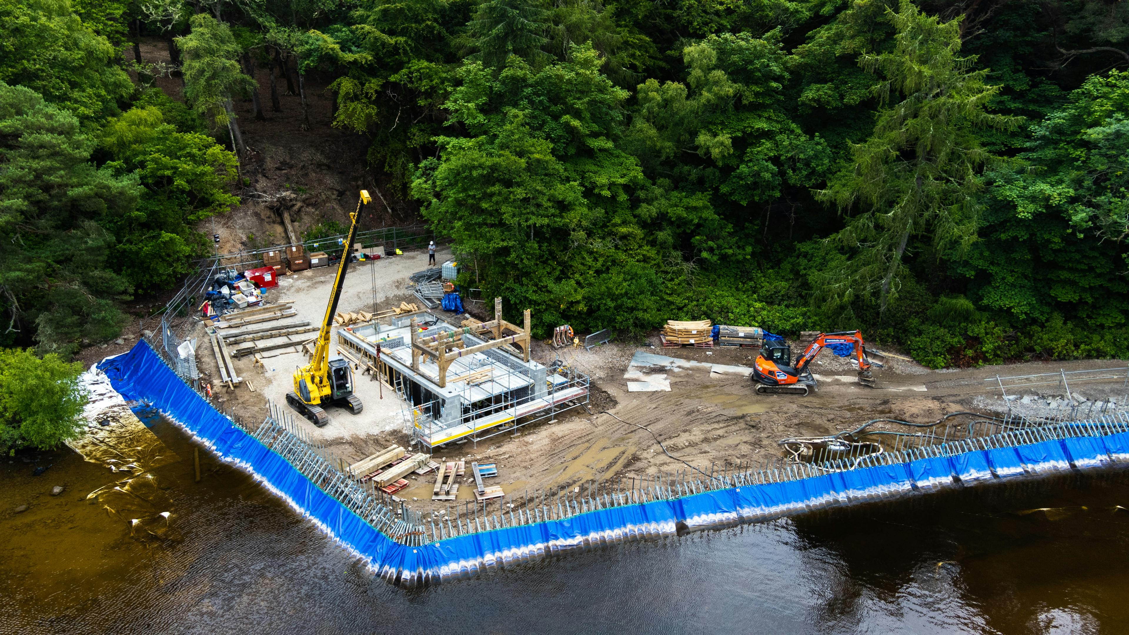 A view of a lake and surrounding green trees and hills with a lake side construction site for a timber framed boathouse in the foreground