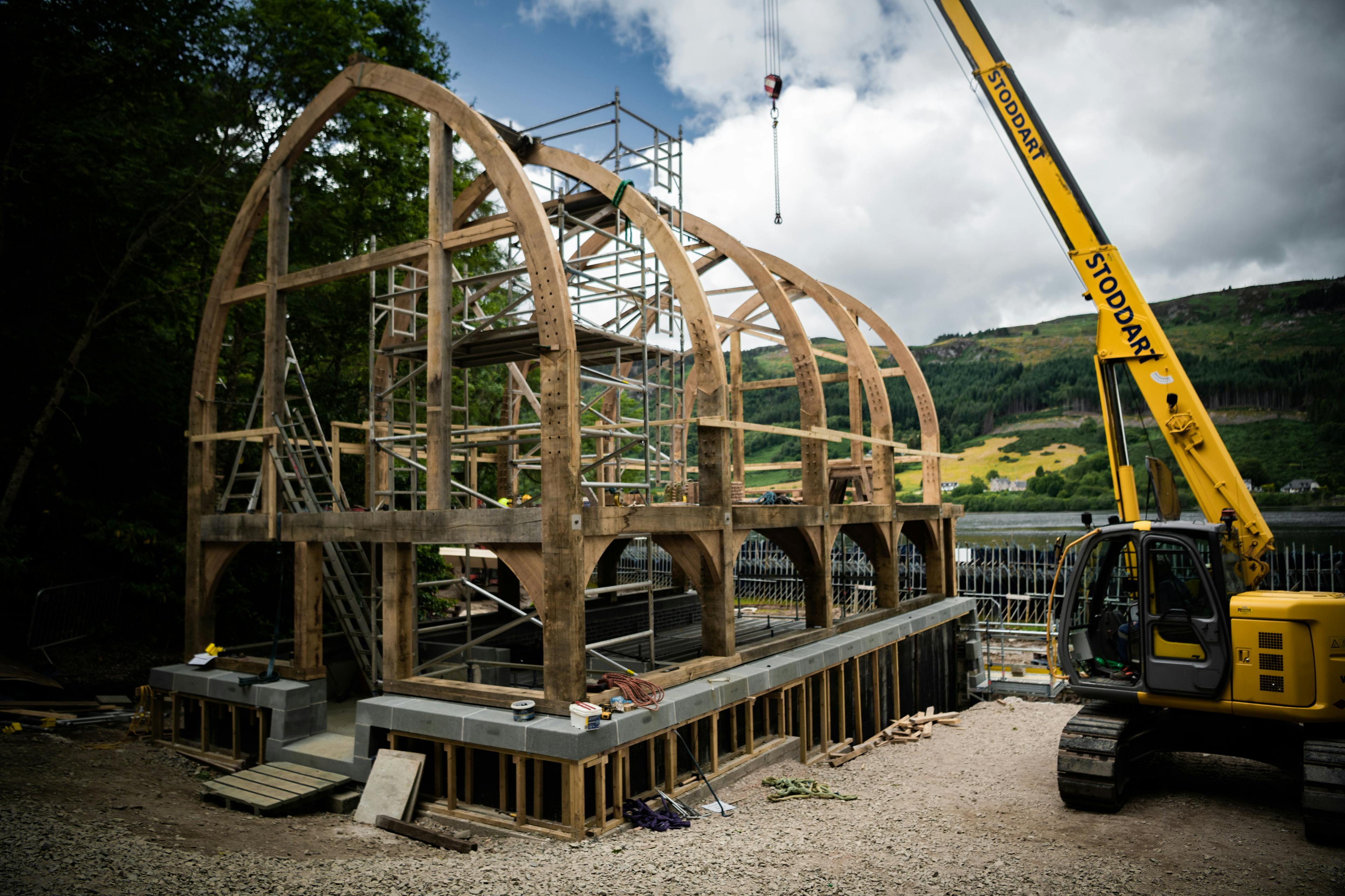 A yellow crane lifts into place the oak frame of a timber boathouse on loch ness with a curved roof during construction