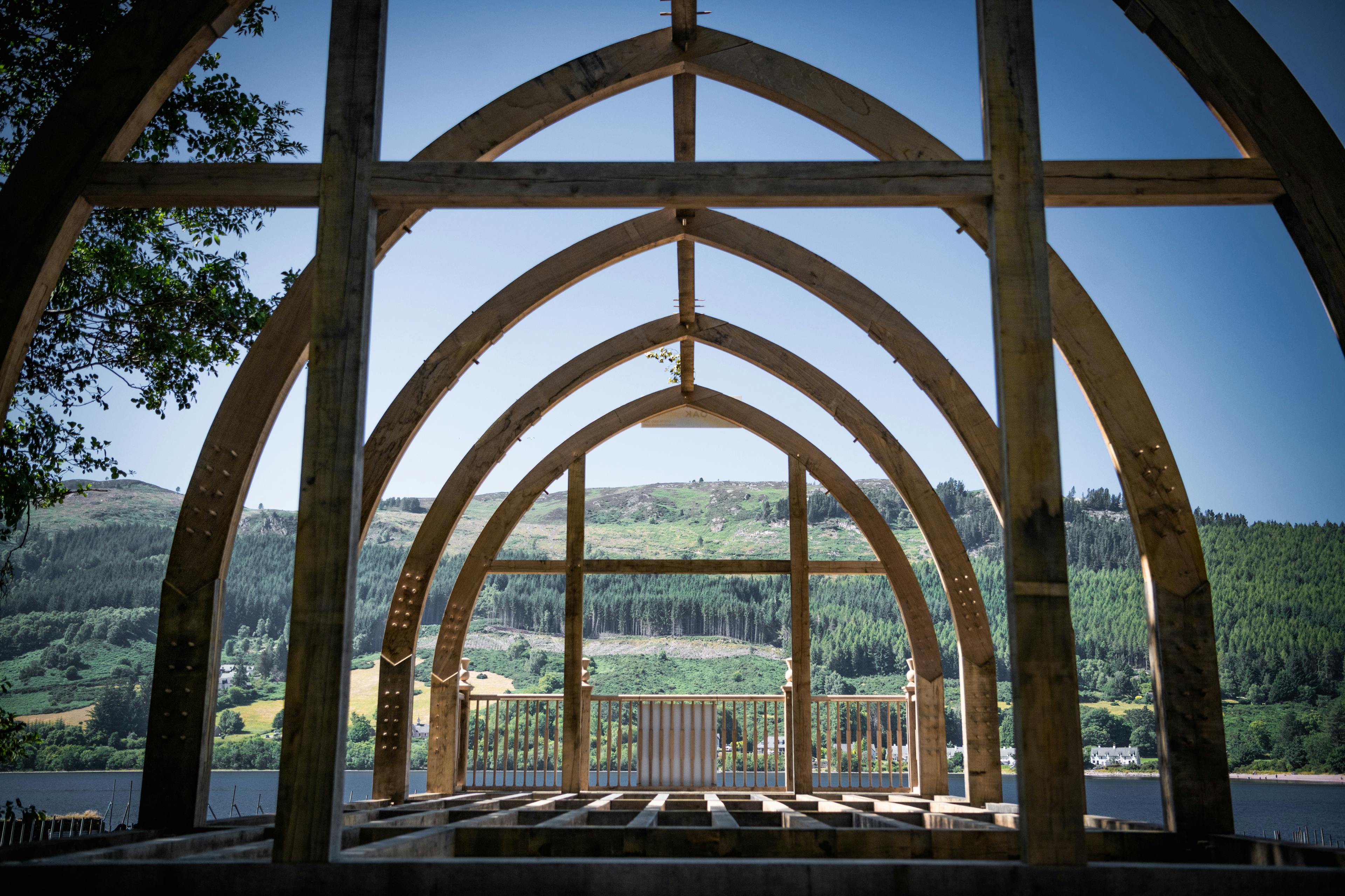 A blue sky and the other side of the loch show through the oak frame of a timber boathouse on loch ness with a curved roof during construction