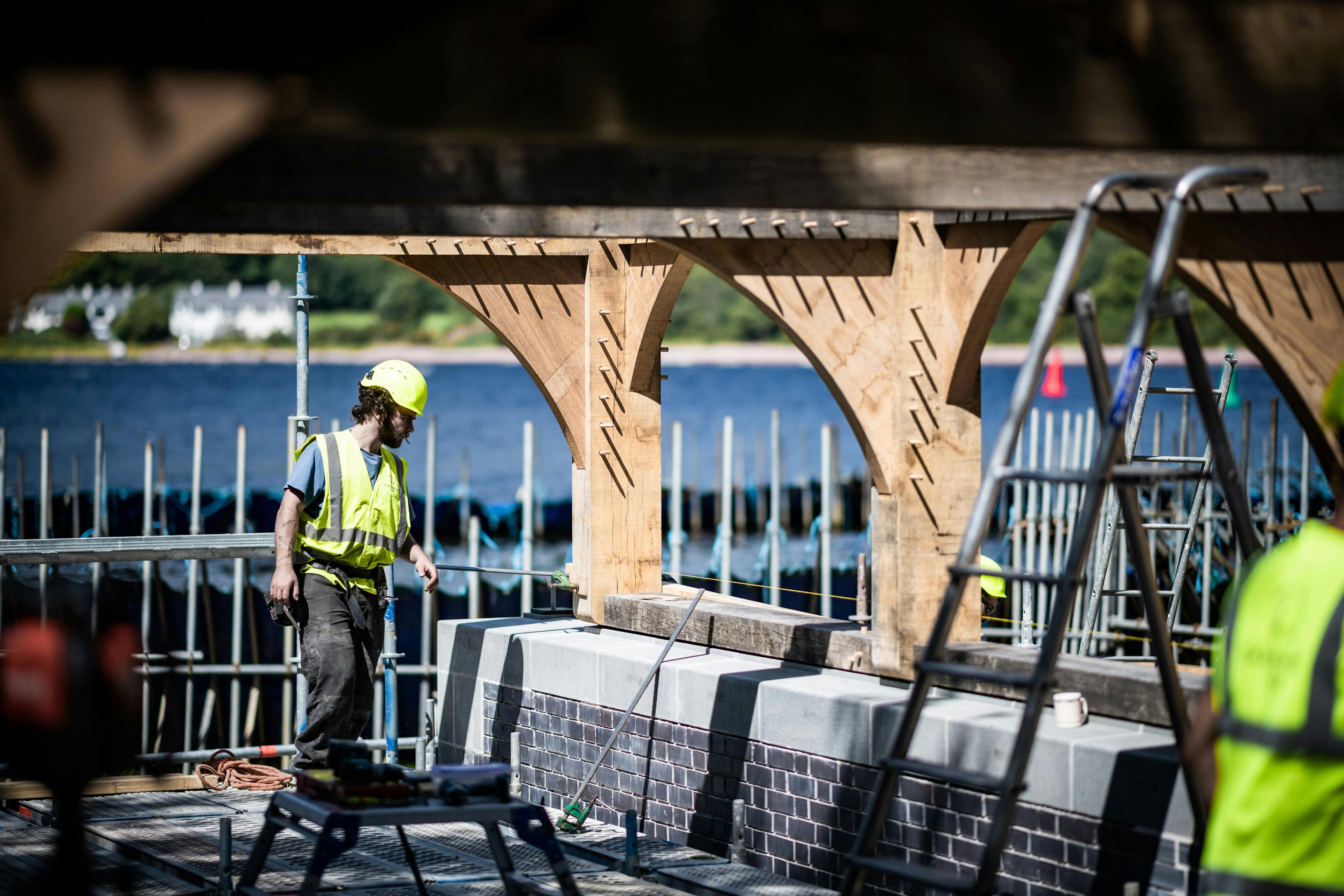 Carpenters in high vis jackets and hard hats install an oak framed boathouse with a curved roof by a lake