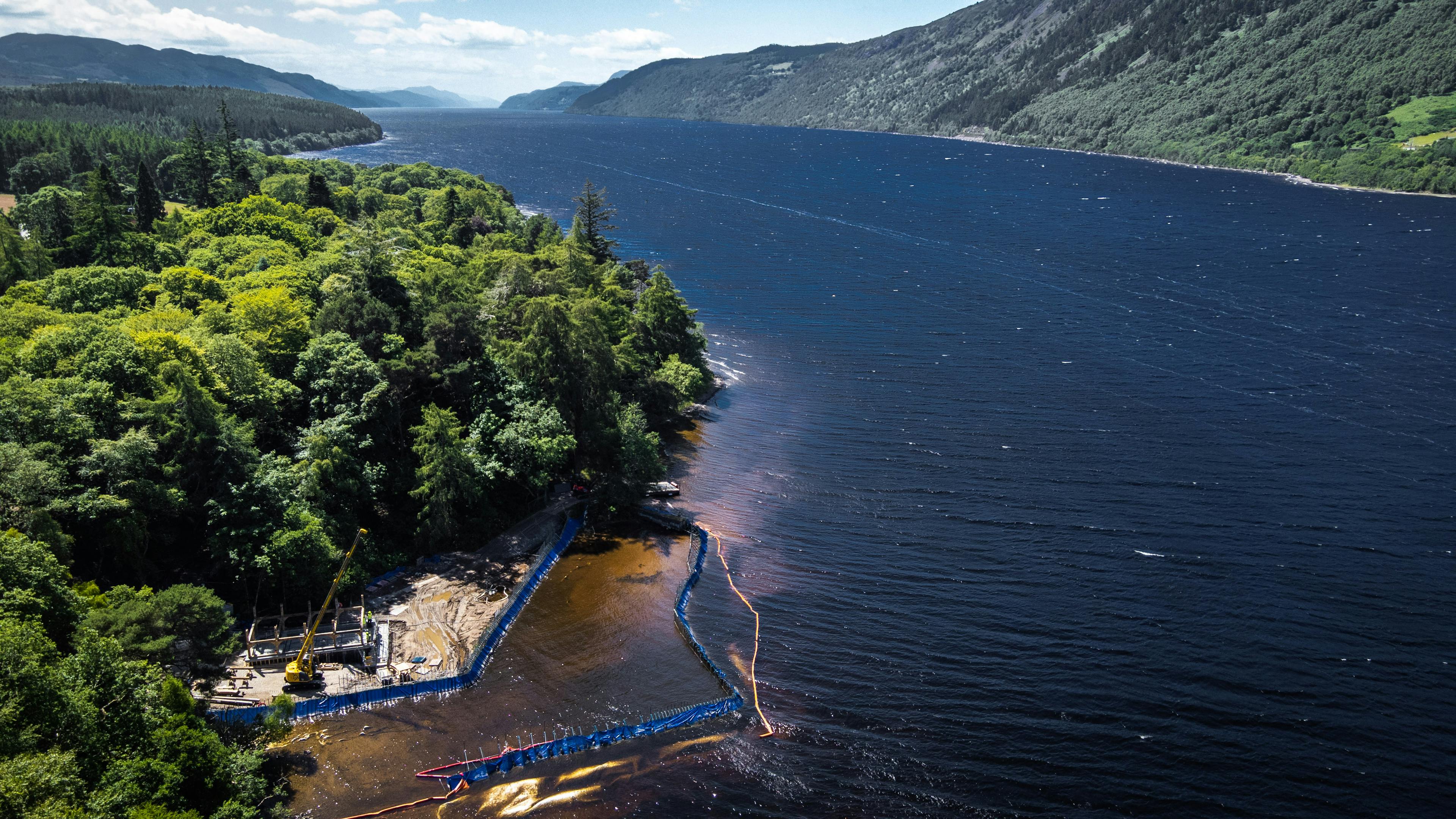 A view of a lake and surrounding green trees and hills with a lake side construction site for a timber framed boathouse in the foreground 