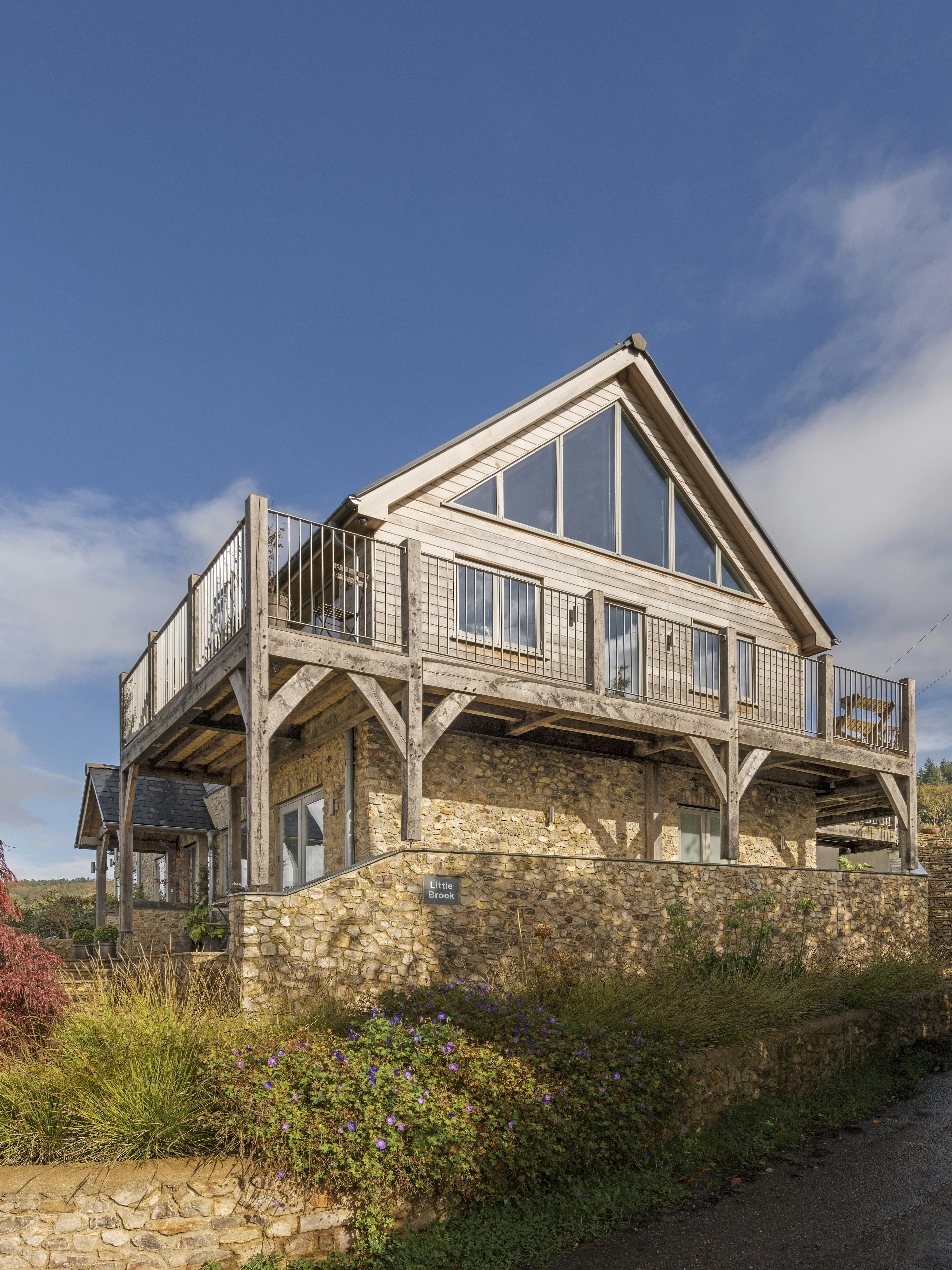 A blue sky behind a timber clad oak framed house with oak balconies wrapping round, and a garden and paved patios in front