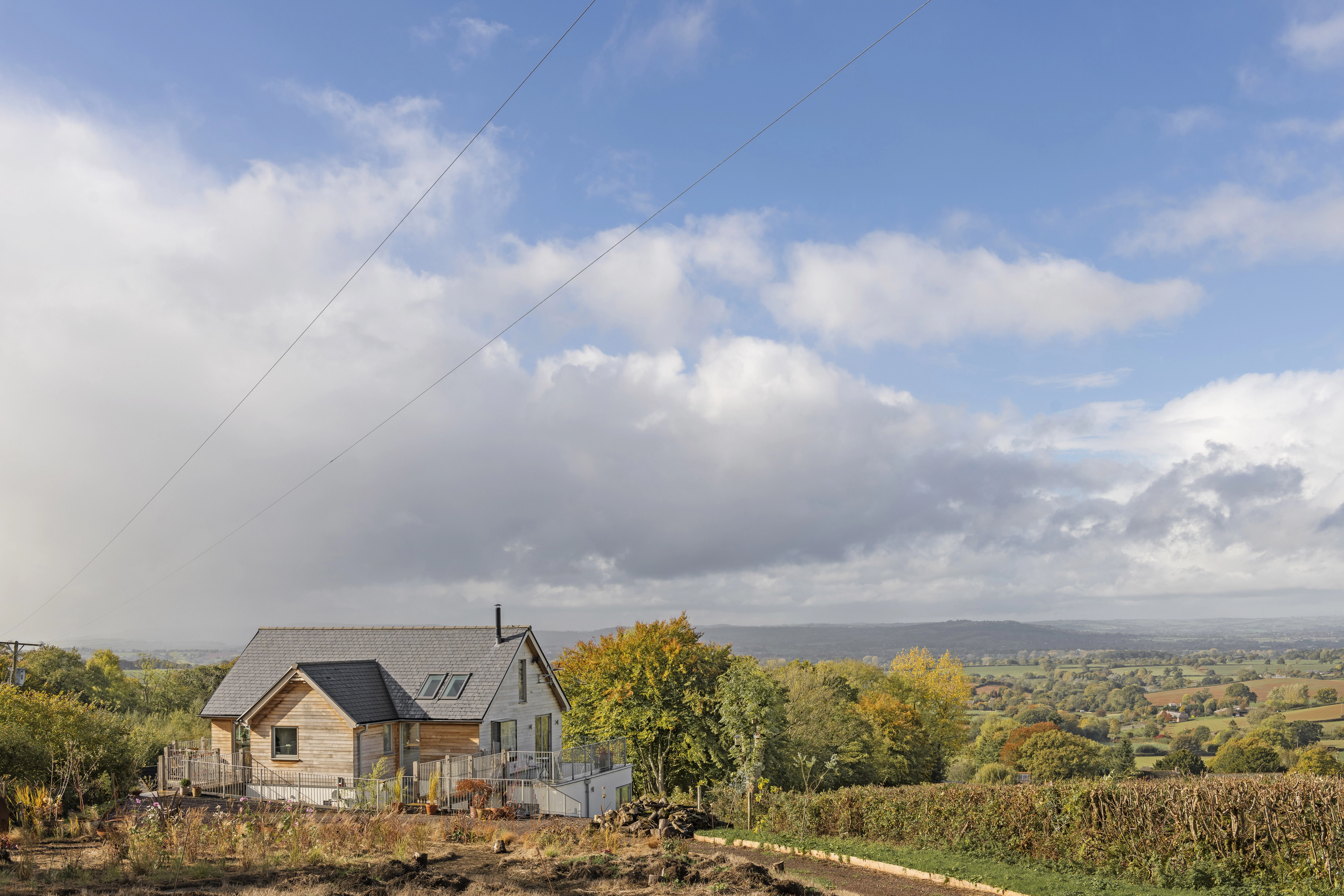 White clouds and a blue sky behind a timber clad oak framed house with oak balconies wrapping round, with green fields and hills in the distance
