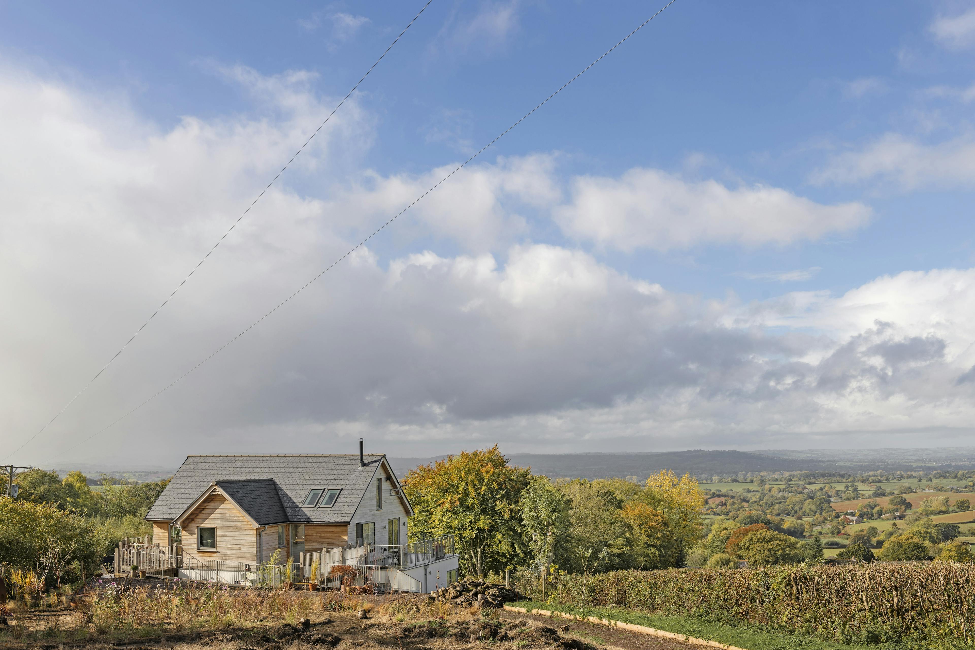 White clouds and a blue sky behind a timber clad oak framed house with oak balconies wrapping round, with green fields and hills in the distance