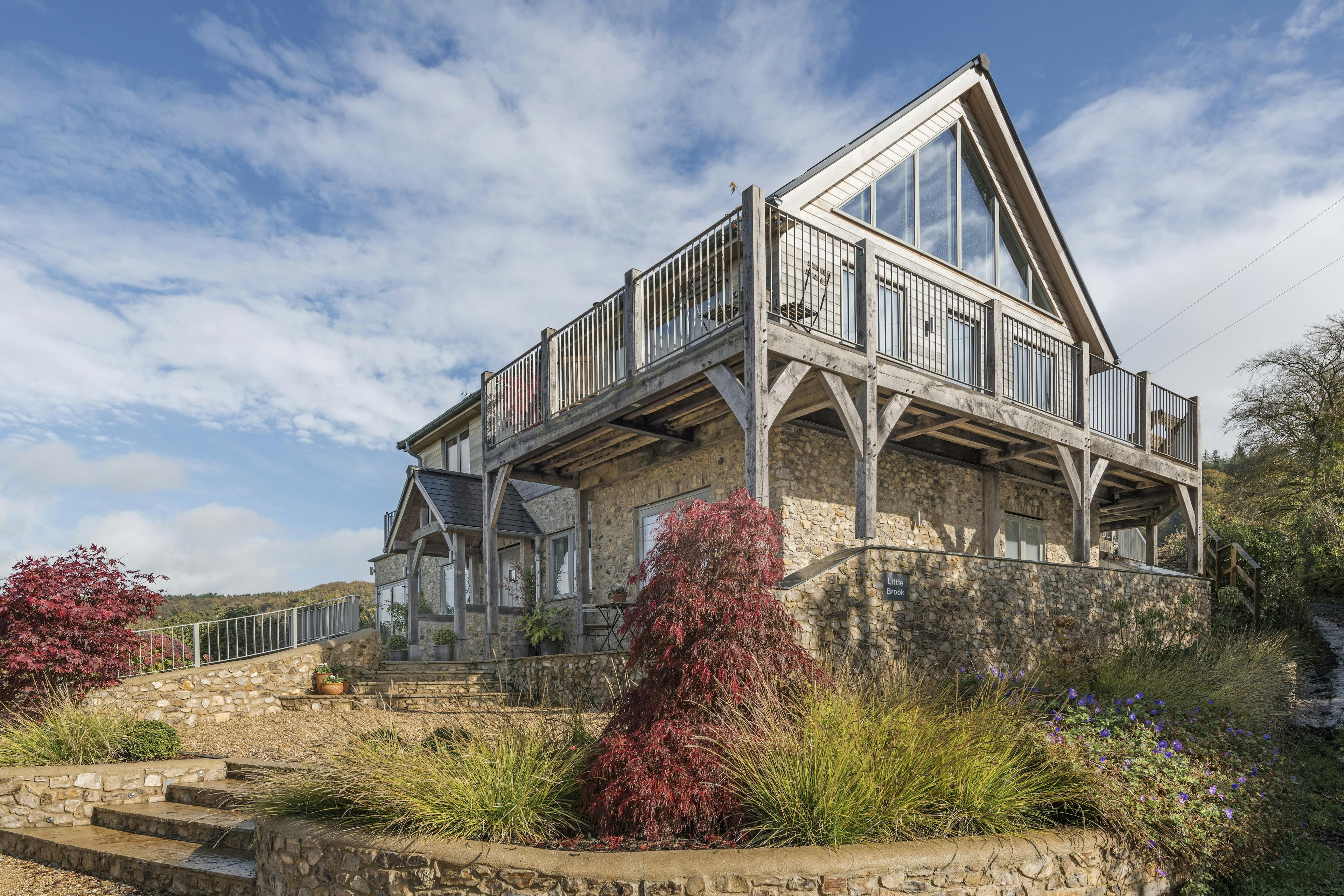 White clouds and a blue sky behind a timber clad oak framed house with oak balconies wrapping round, and a garden and paved patios in front