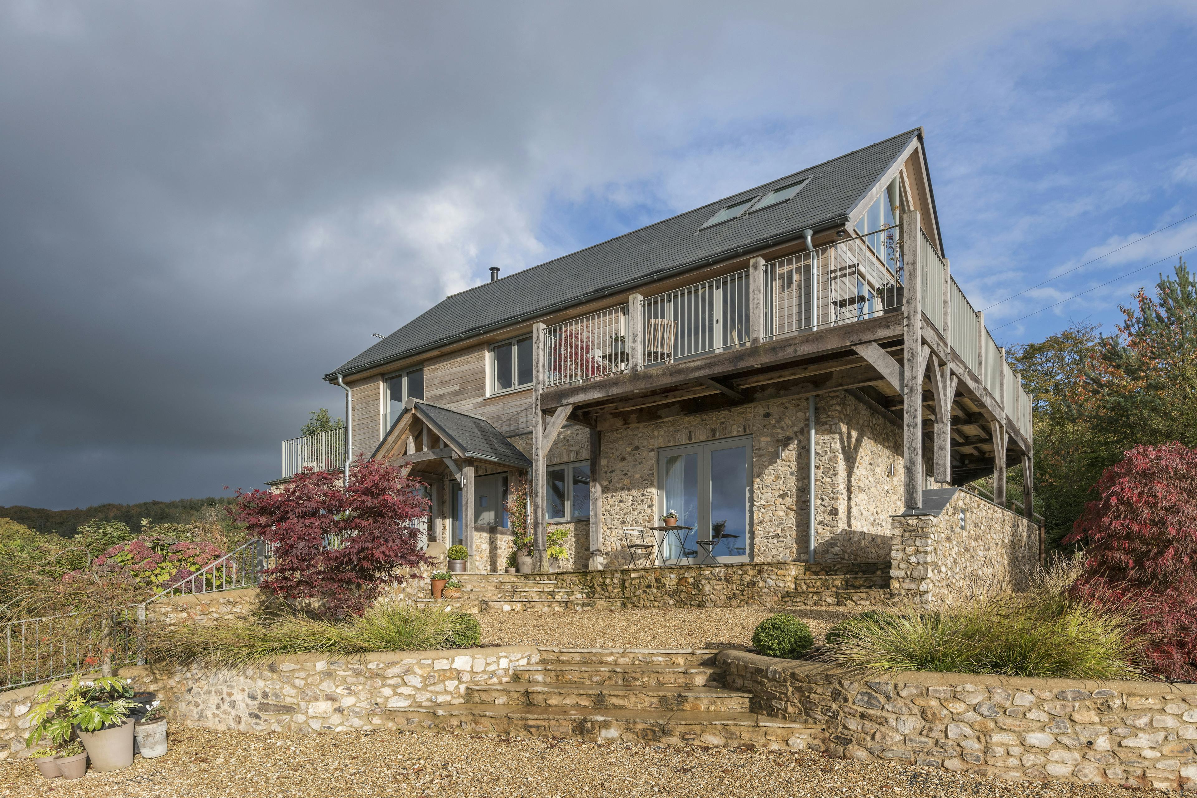 Dark clouds behind a timber clad oak framed house with oak balconies wrapping round, and a garden and paved patios in front