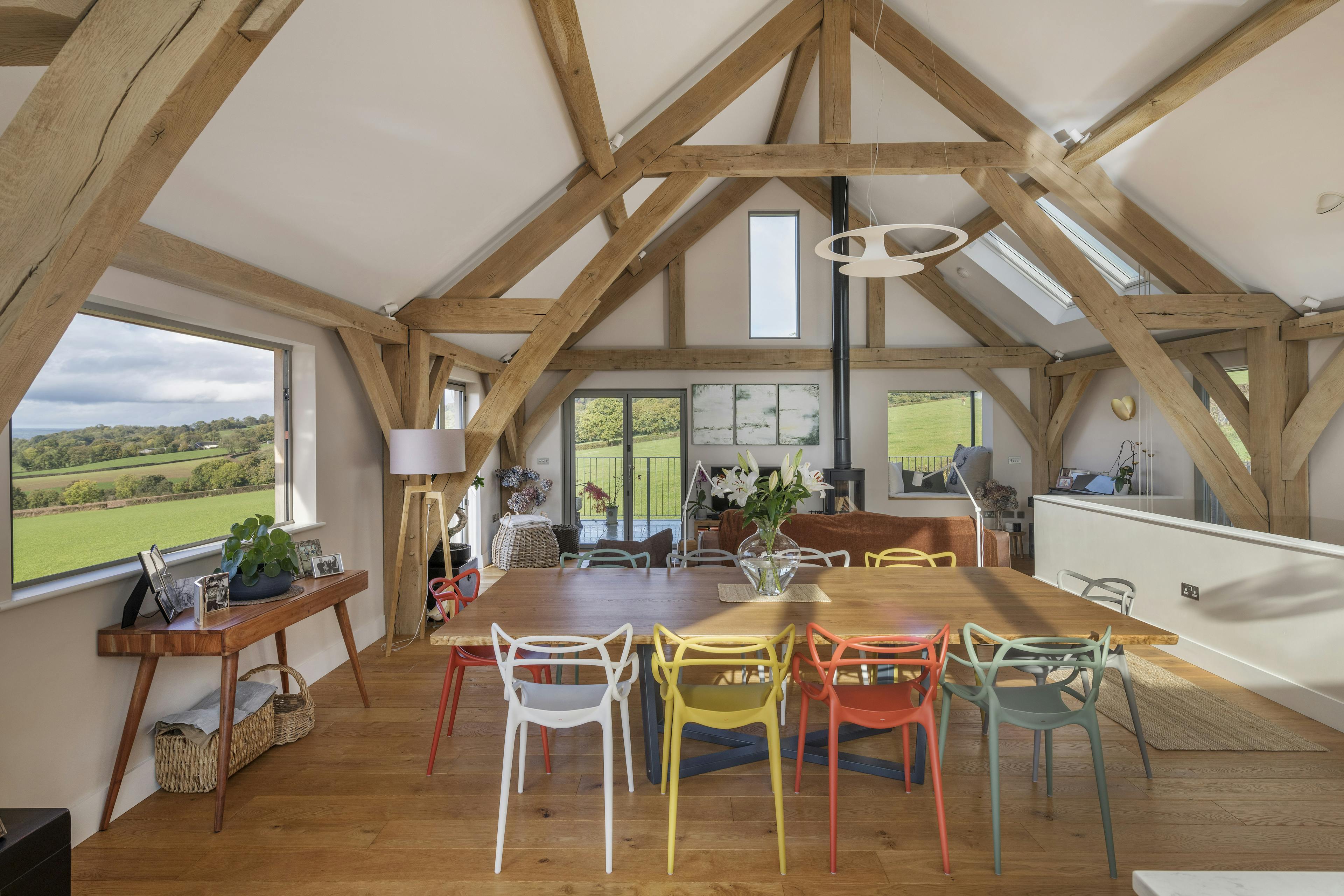 A dining table, sitting area and log burner in a vaulted oak framed open plan living room with a large rectangle bi fold window opens out to views of green countryside