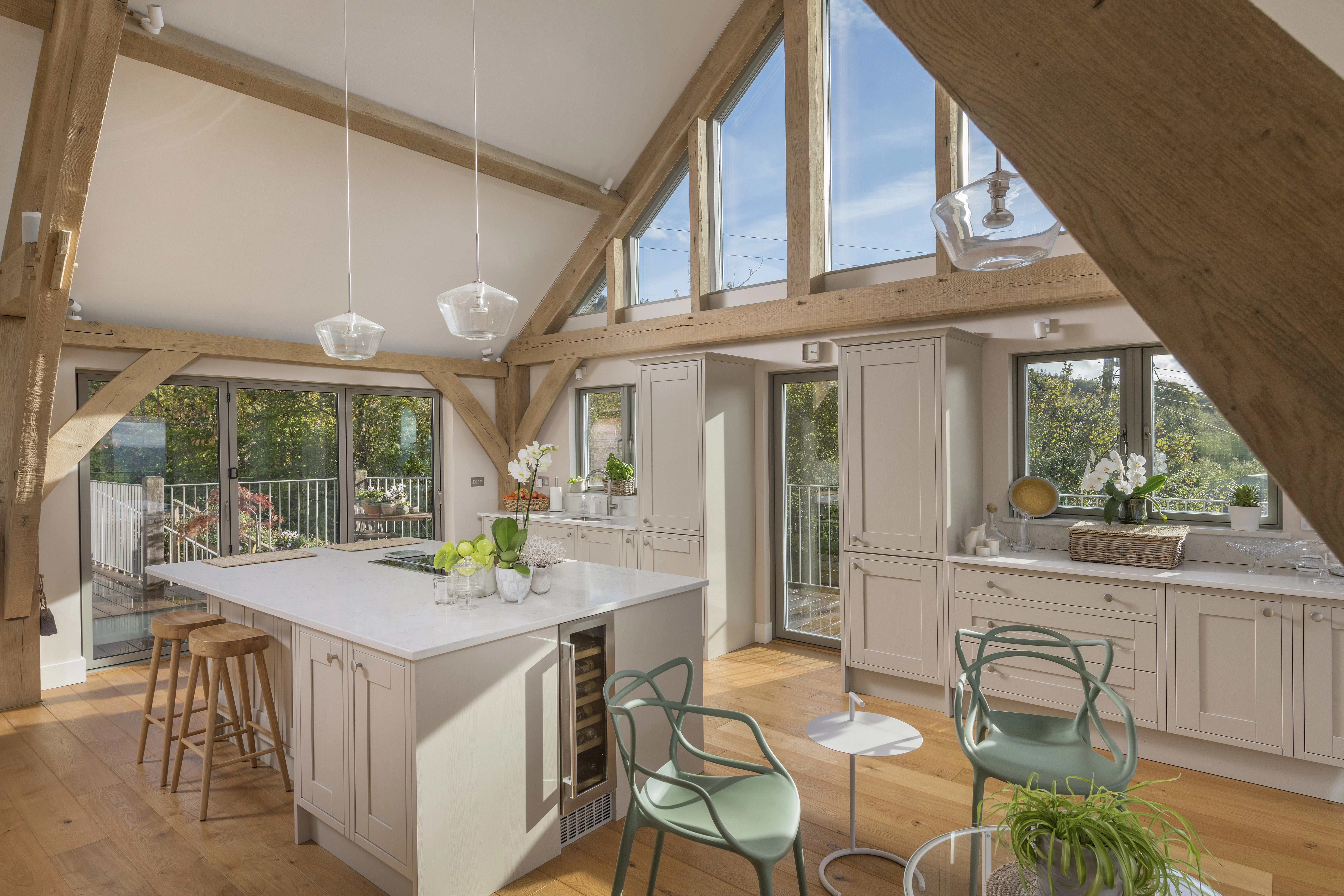 Sunlight beams through glazed windows in a kitchen with a large island and small table and chairs in a vaulted oak framed open plan living room