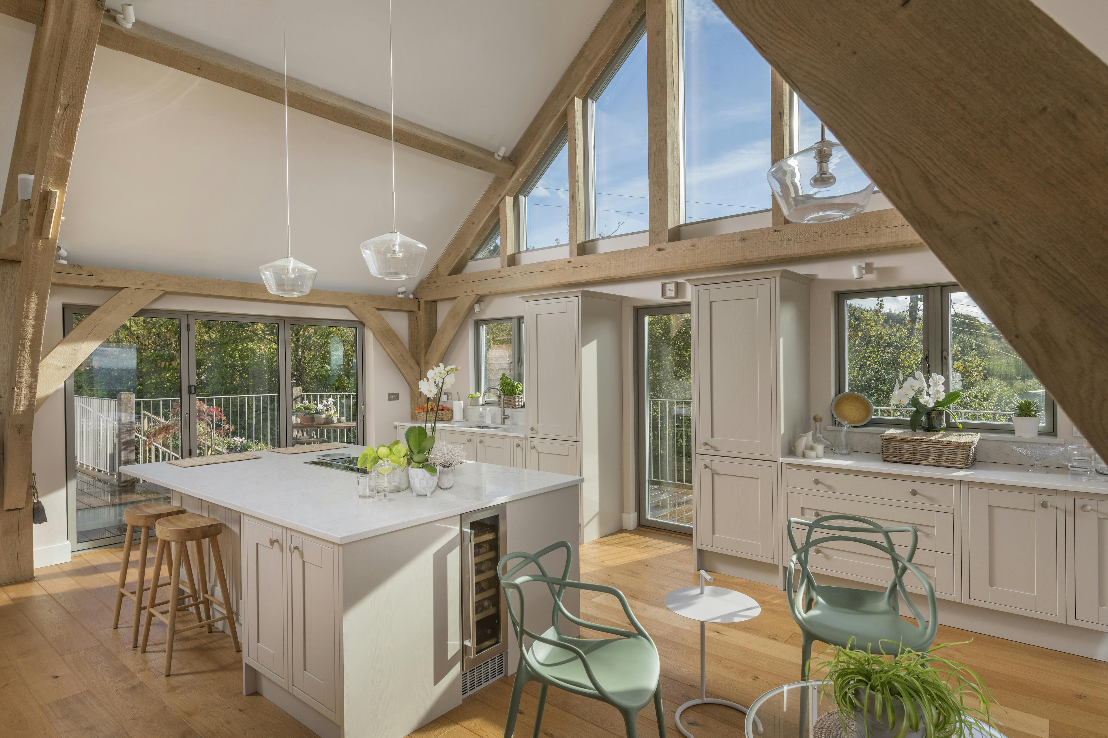 Sunlight beams through glazed windows in a kitchen with a large island and small table and chairs in a vaulted oak framed open plan living room