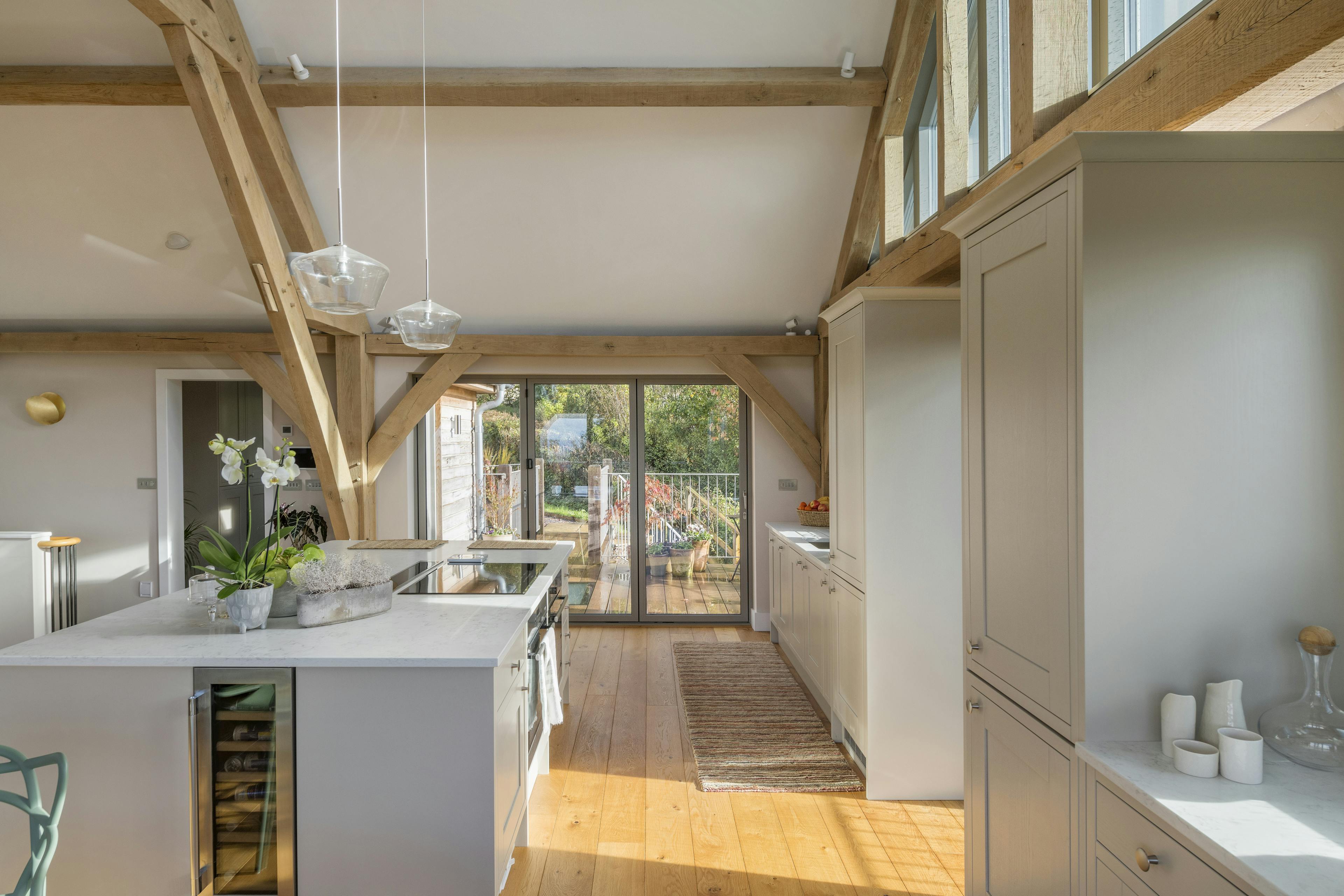 Sunlight beams through glazed windows in a kitchen with a large island in a vaulted oak framed open plan living room