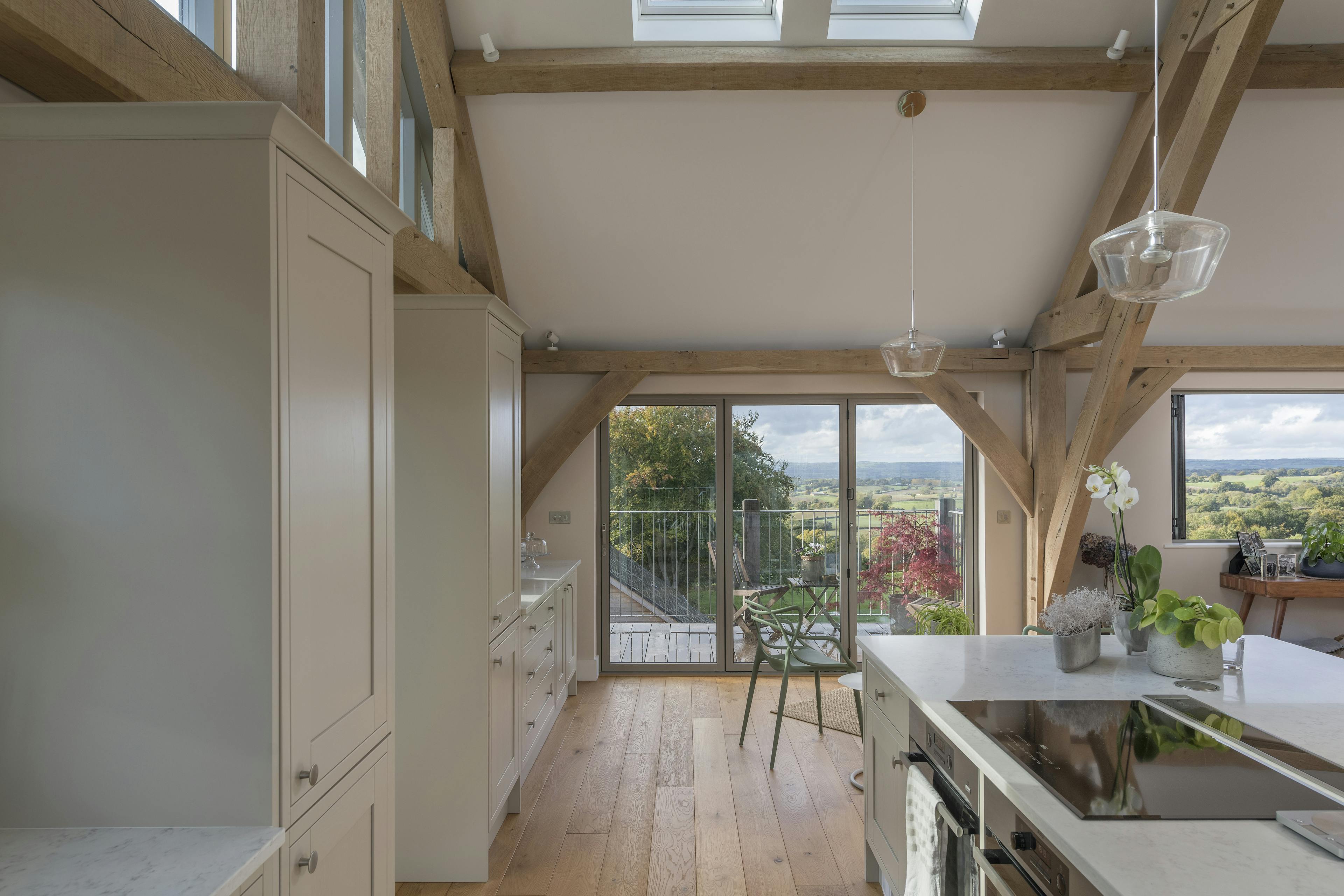 A kitchen with a large island and small table and chairs in a vaulted oak framed open plan living room with glazed bi fold doors and views of the green countryside