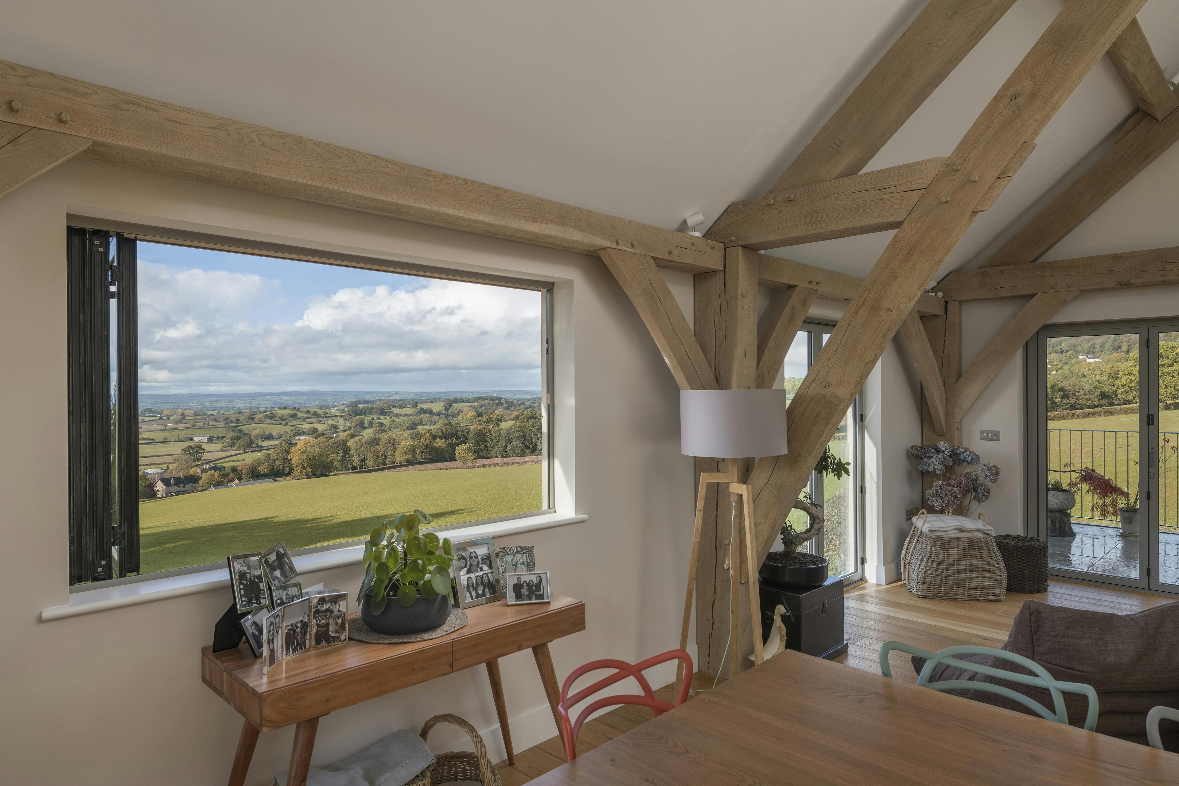A large rectangle bi fold window opens out to views of green countryside in a vaulted oak framed open plan living room