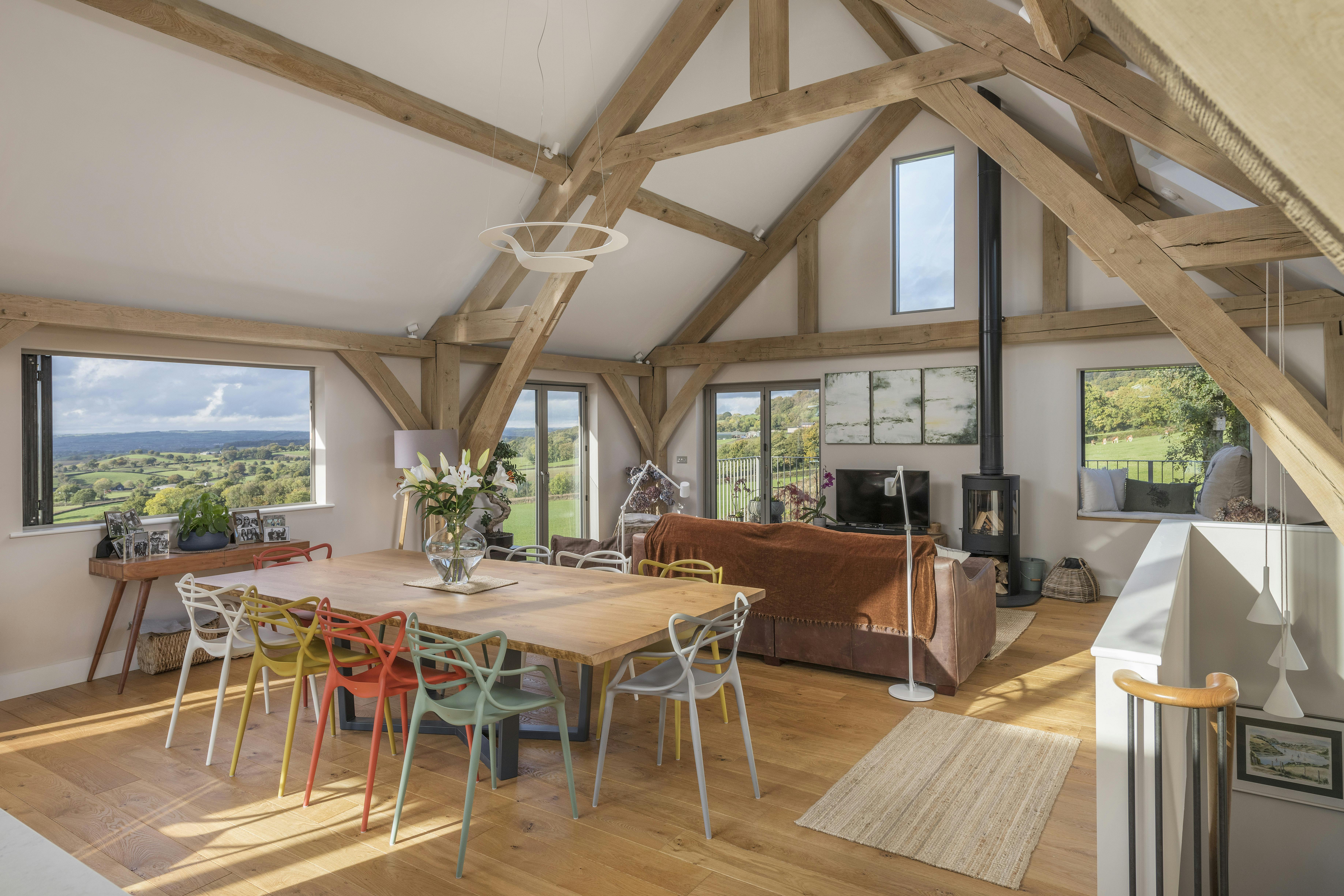A dining table, sitting area and log burner in a vaulted oak framed open plan living room with a large rectangle bi fold window opens out to views of green countryside