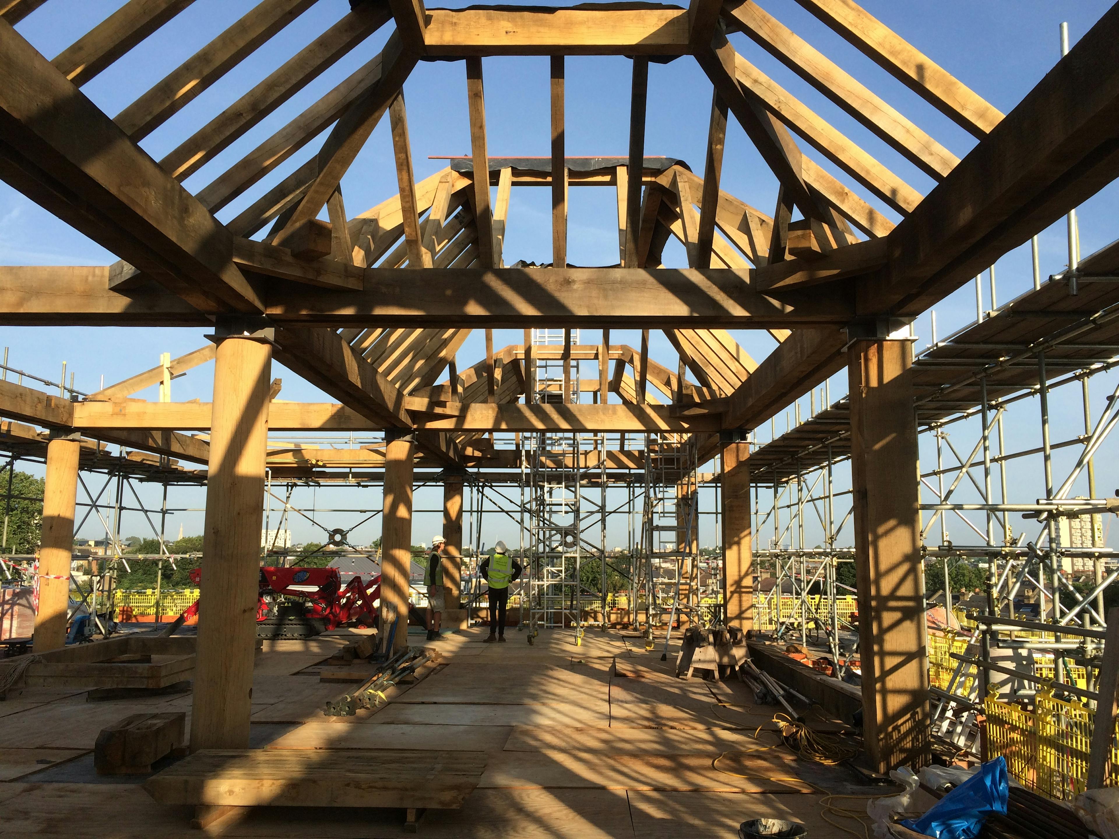 Turned oak columns hold up oak lanterns on a construction site for a rooftop bar in a city with blue skies showing between the timber beams