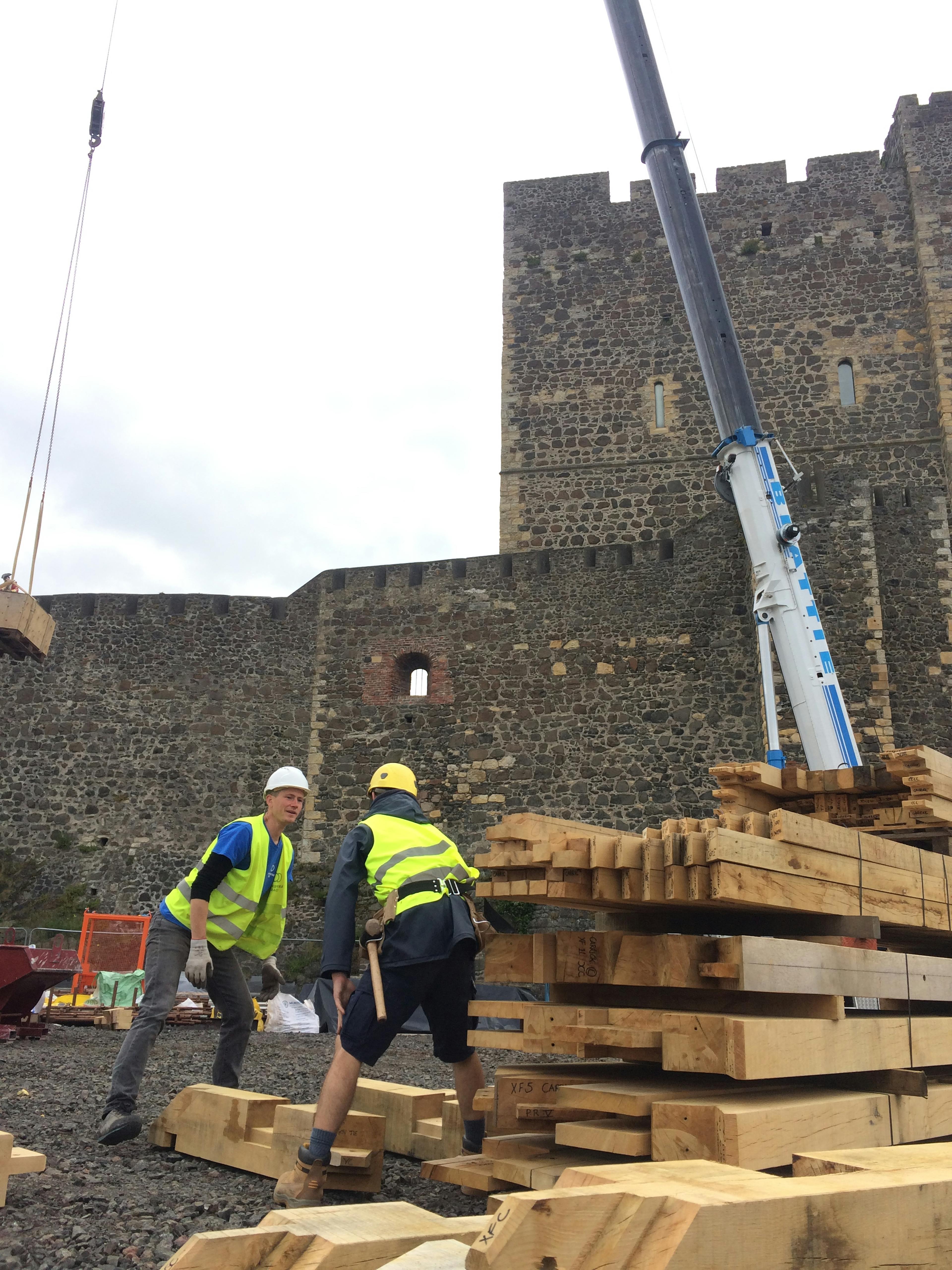 On a construction site by a stone castle, carpenters with high vis jackets oversee an oak frame which is stacked and ready to be lifted by a crane 