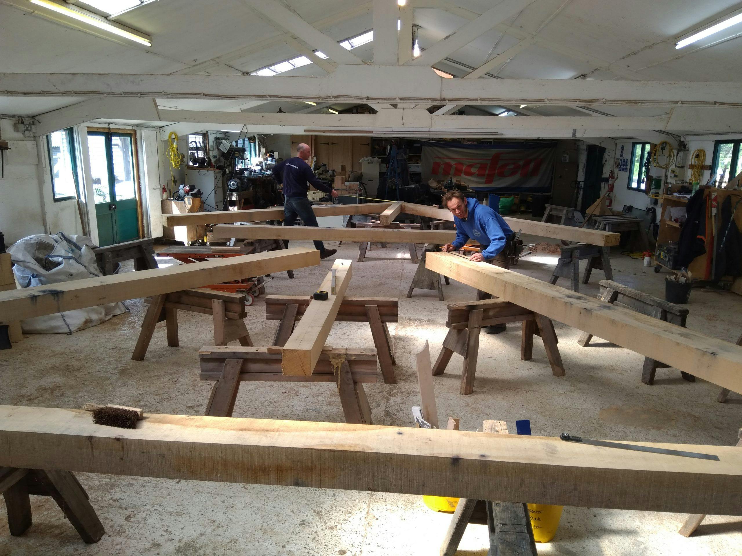 An oak frame laid out on trestle tables in a workshop with carpenters working with tools on the frame