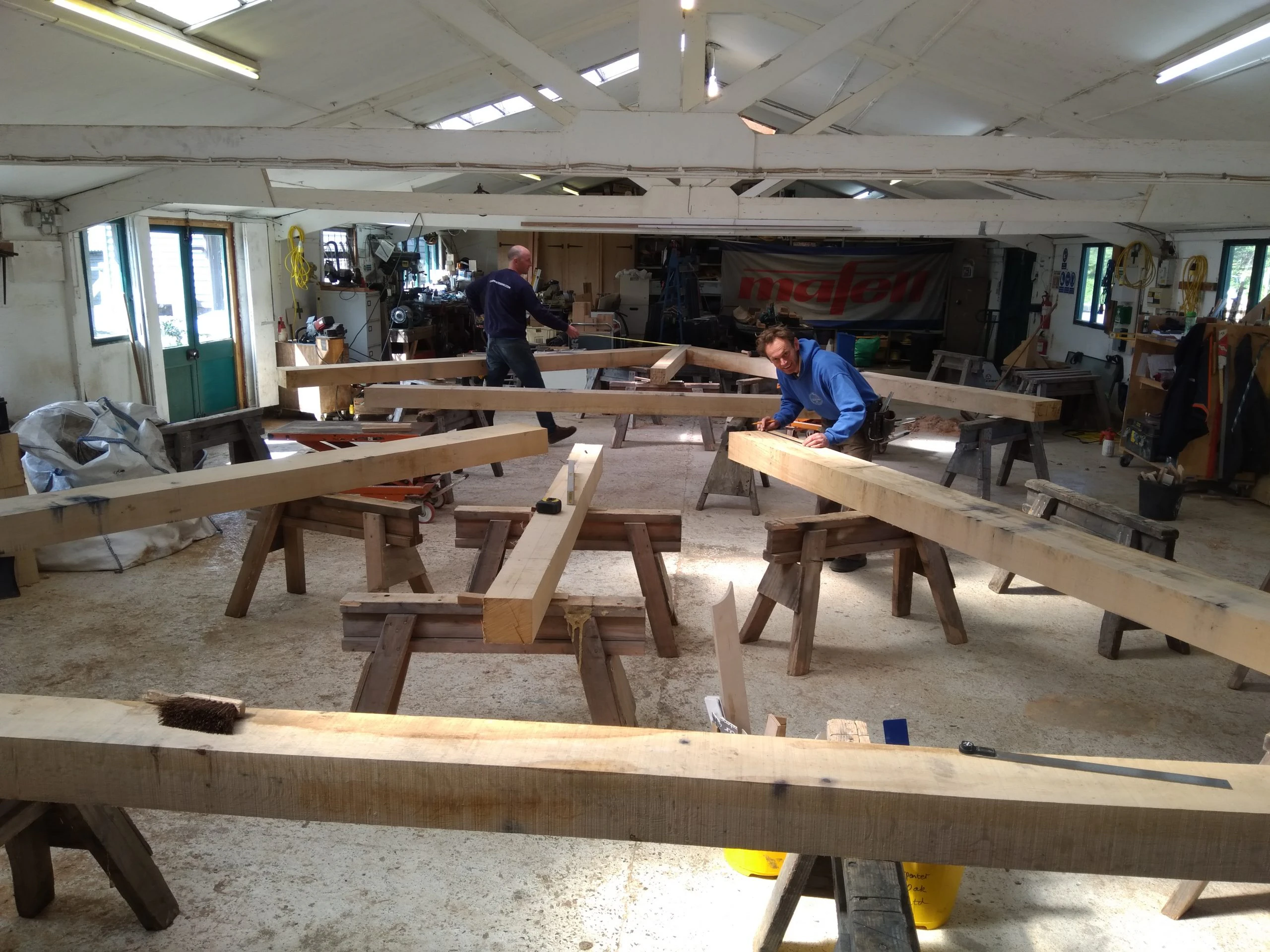 An oak frame laid out on trestle tables in a workshop with carpenters working with tools on the frame