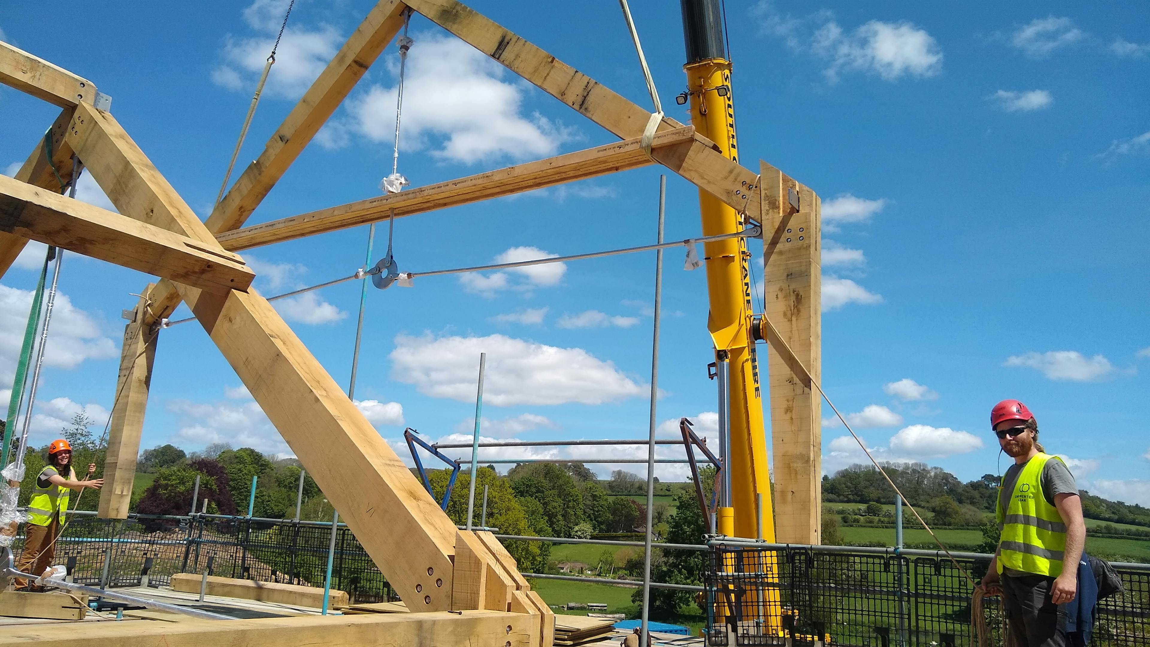The truss of an oak frame is put in place by a crane on a construction site, with carpenters overseeing