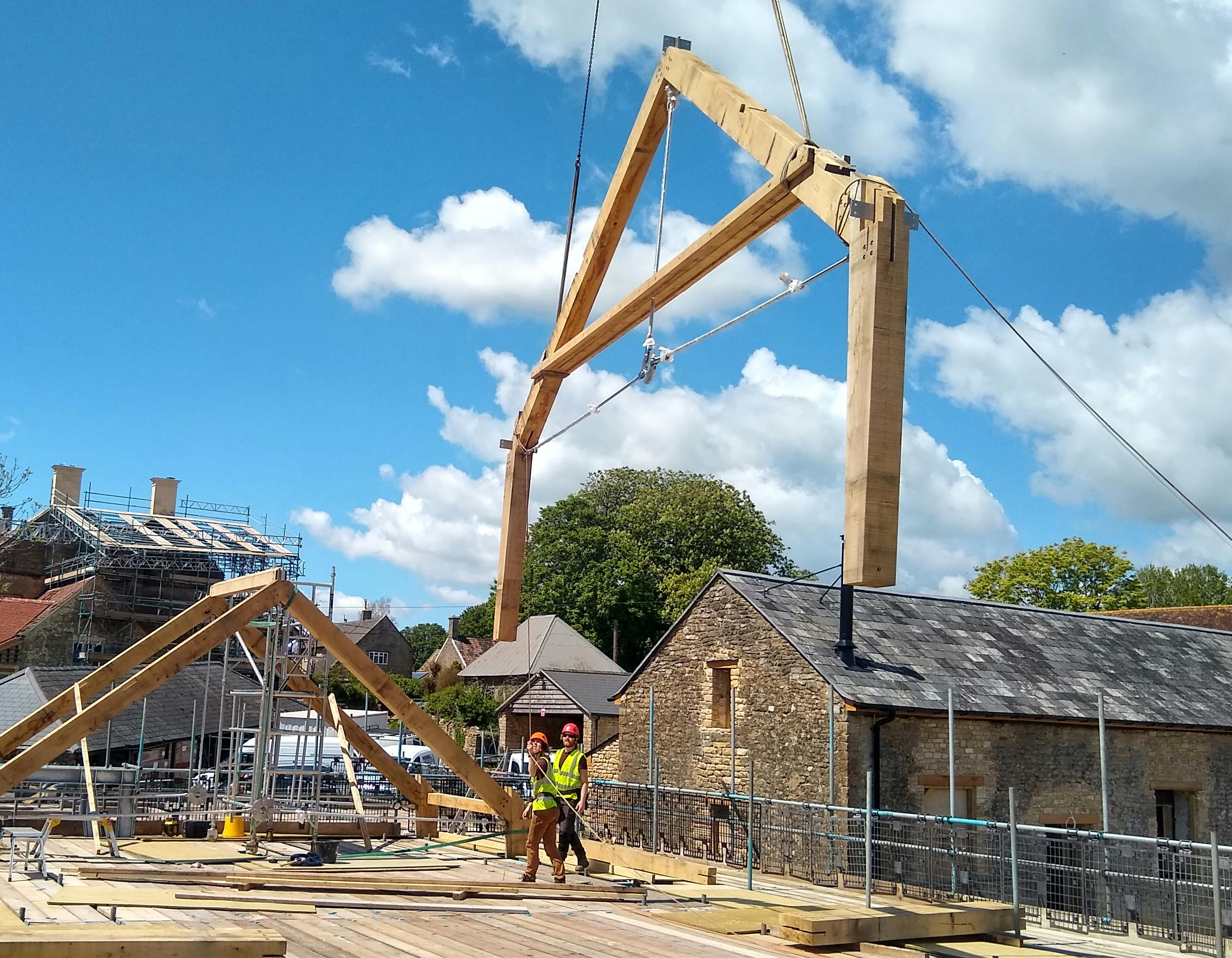 The truss of an oak frame is put in place by a crane on a construction site, with carpenters overseeing