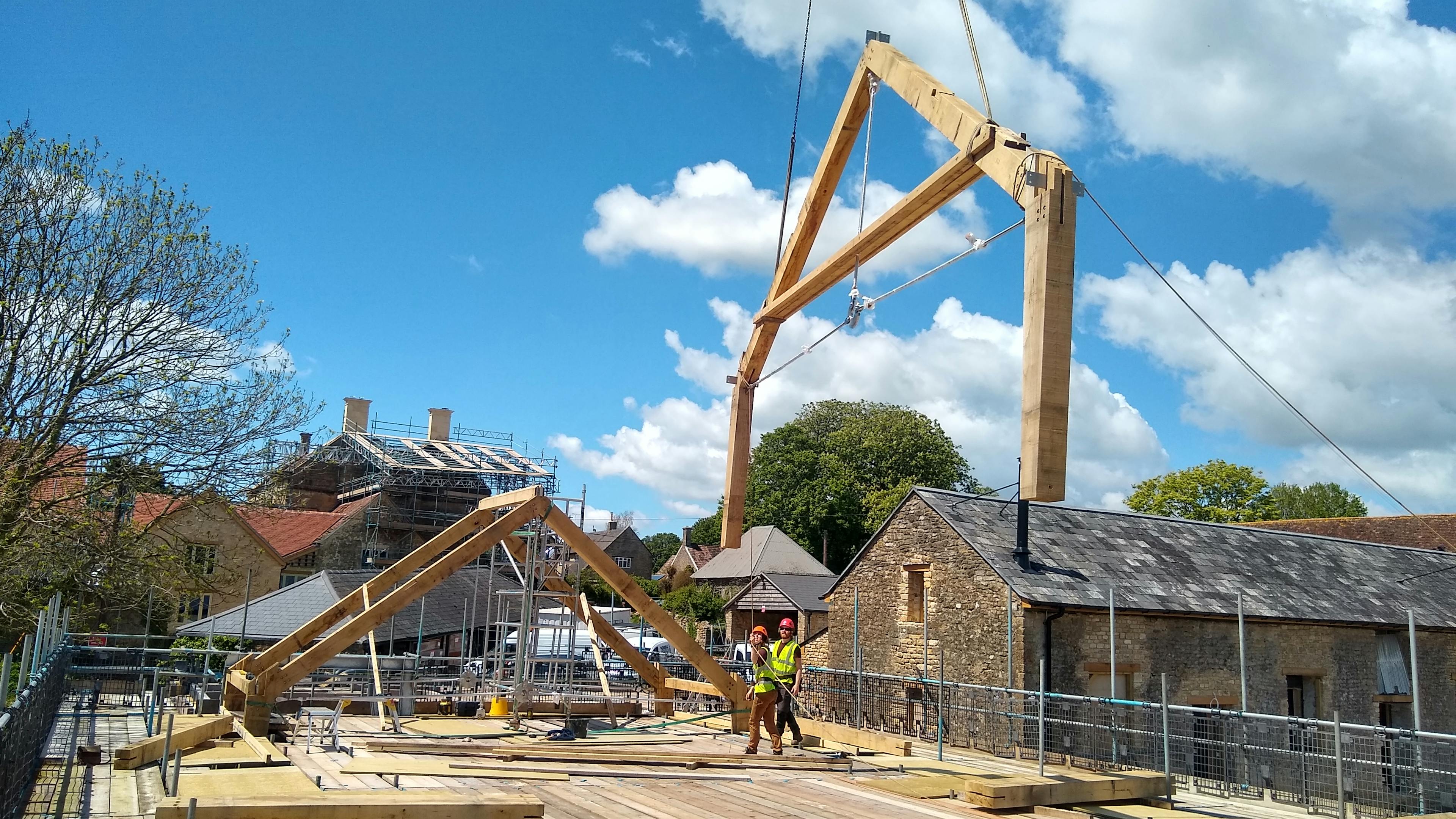The truss of an oak frame is put in place by a crane on a construction site, with carpenters overseeing