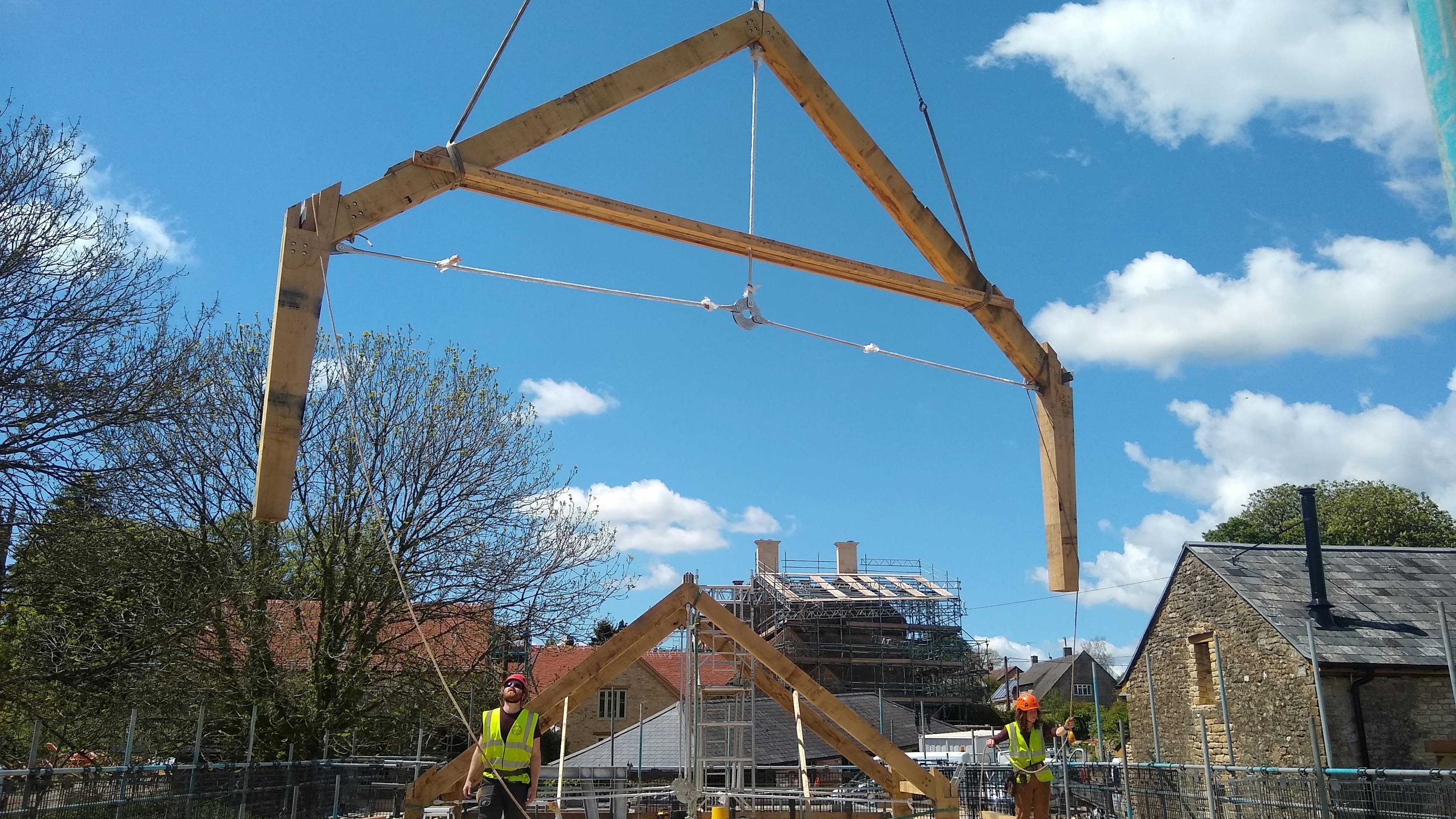 The truss of an oak frame is put in place by a crane on a construction site, with carpenters overseeing
