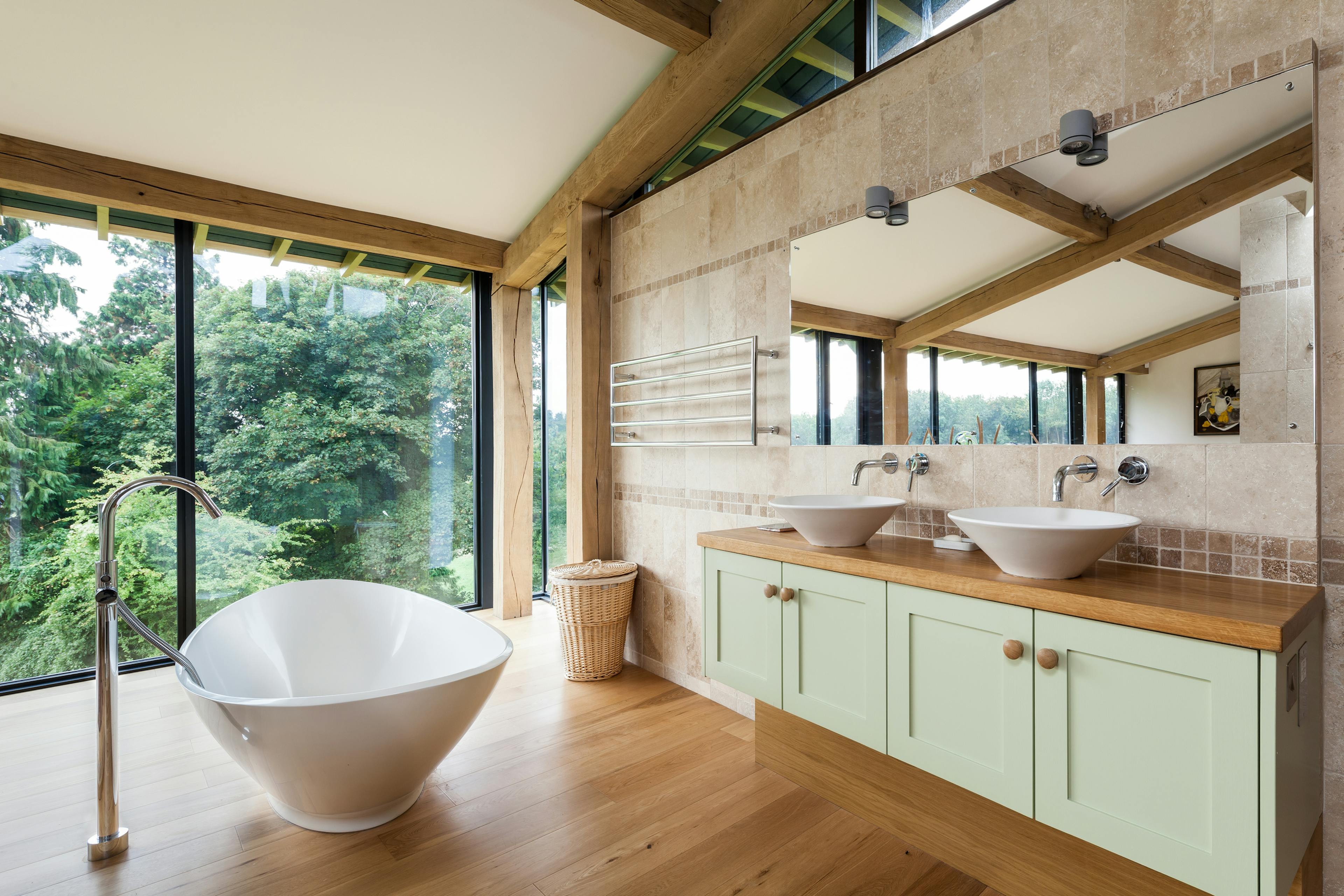 The bathroom of a curved oak framed house with a standalone bath, green cabinets and large glazed windows