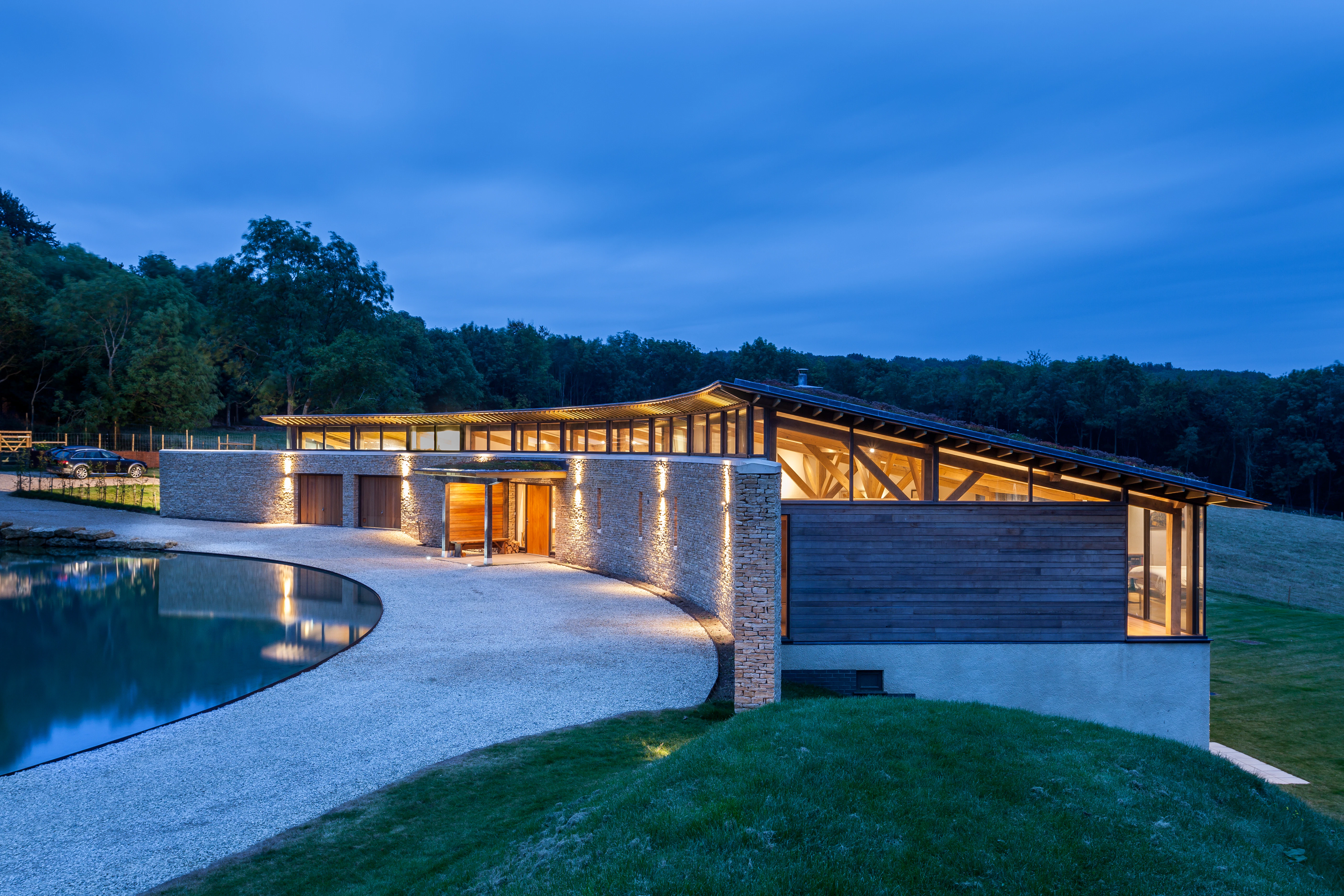 A curved oak framed house with stone finished front wall at dusk with the lights on