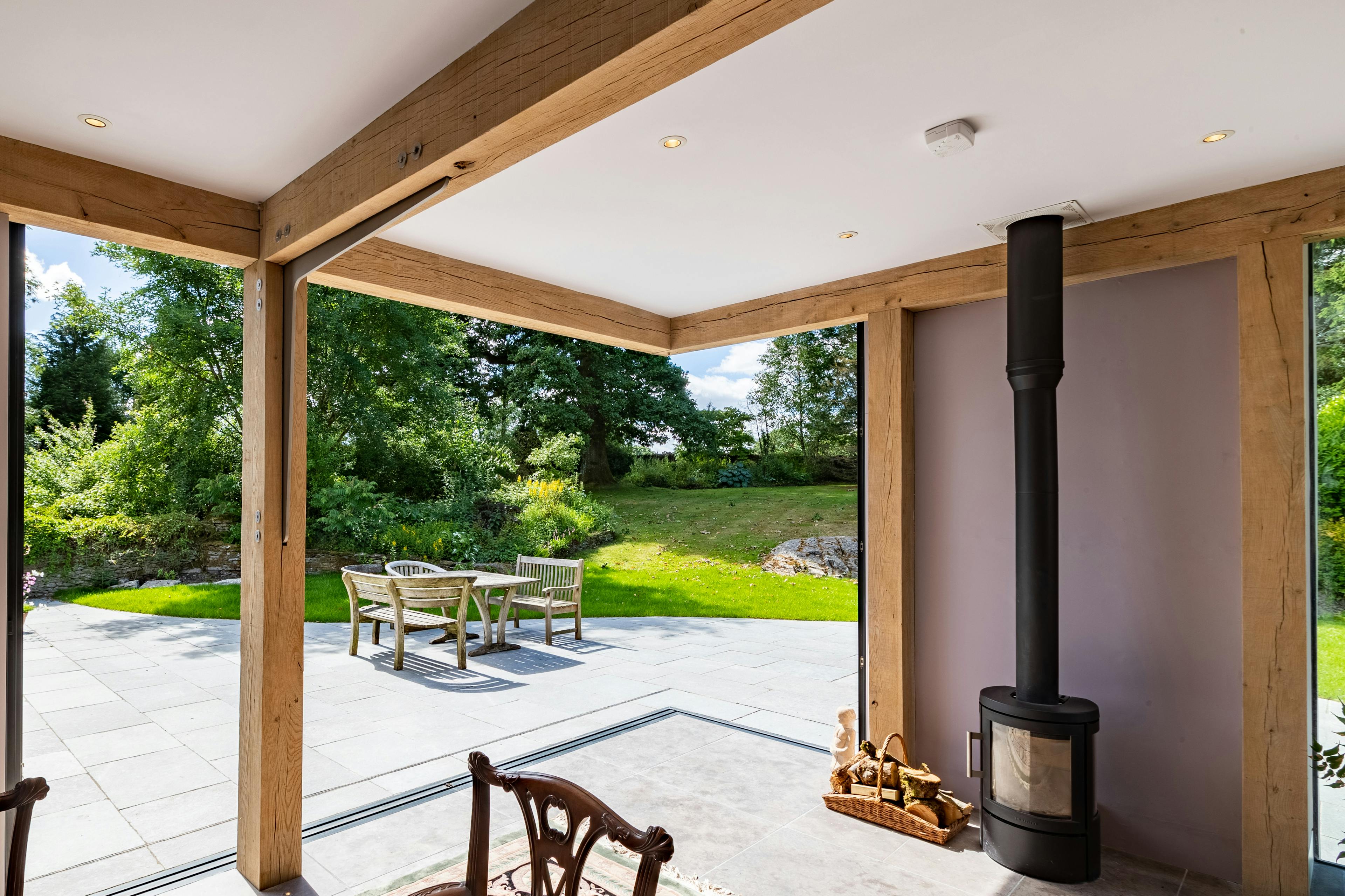 An oak frame living room extension with a log burner, showing a wide opening with direct access to a terrace and garden