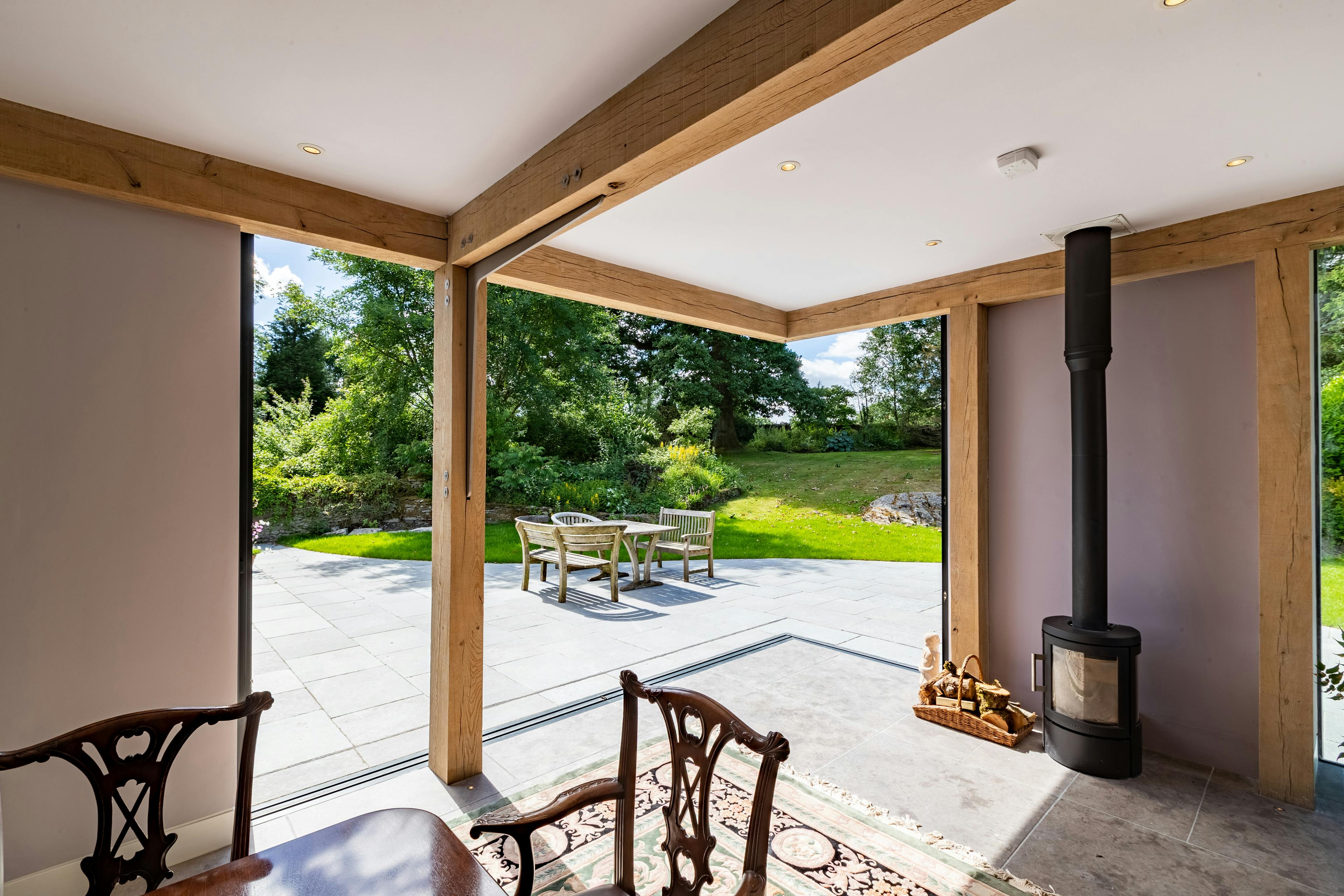 An oak frame living room extension with a log burner, showing a wide opening with direct access to a terrace and garden