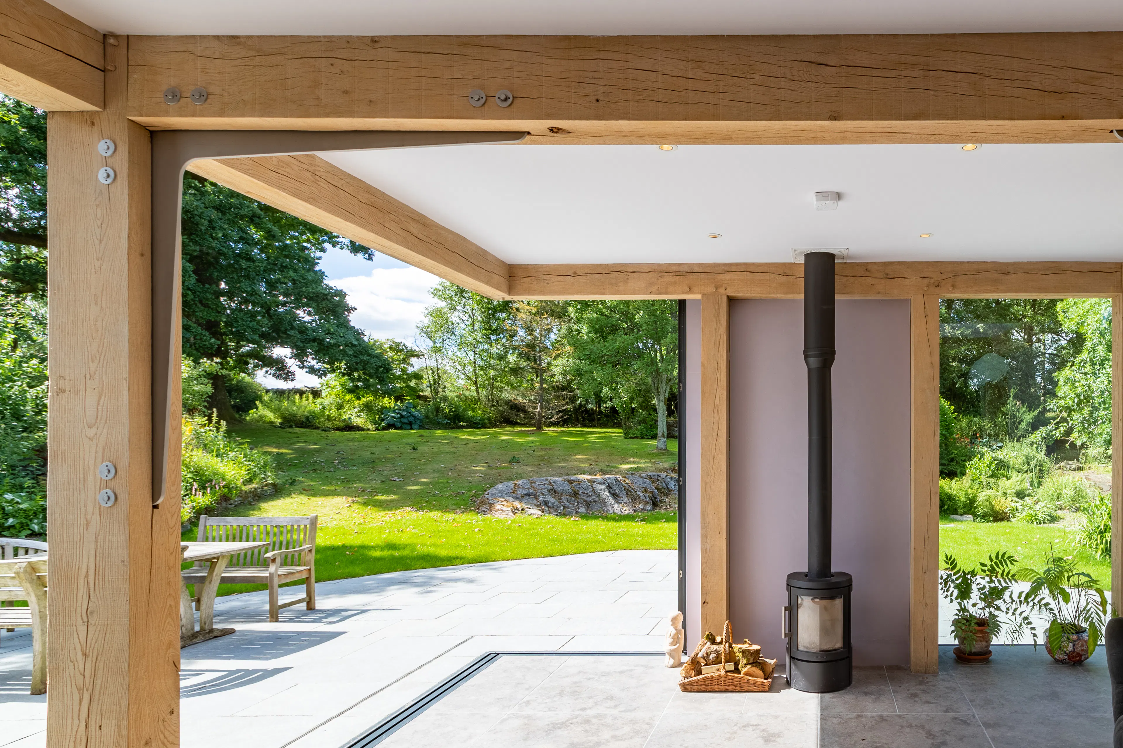 An oak frame living room extension with a log burner, showing a wide opening with direct access to a terrace and garden