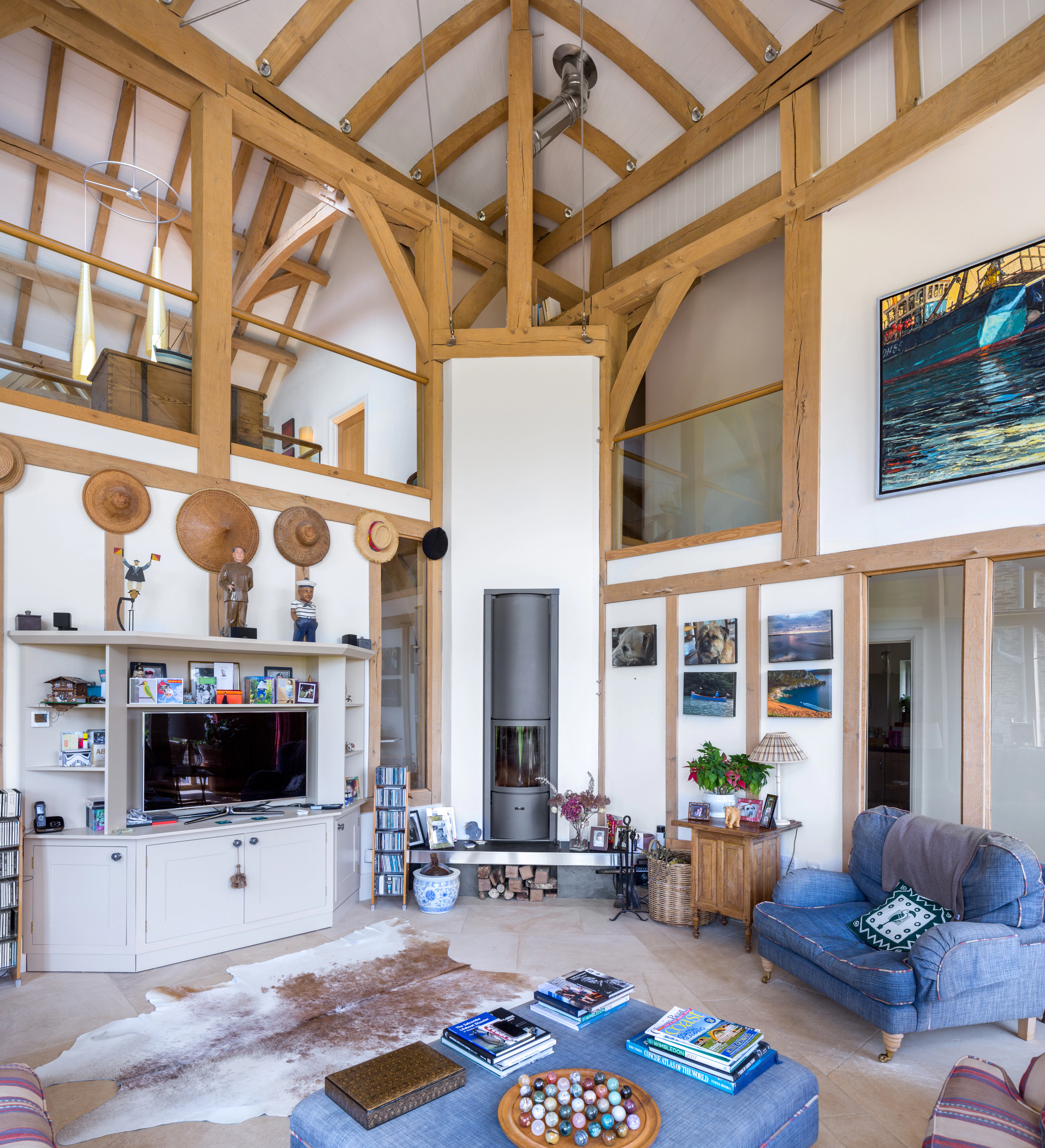 Interior view of a double-height oak-framed family room, showcasing a wrap-around glass mezzanine and a central log burner