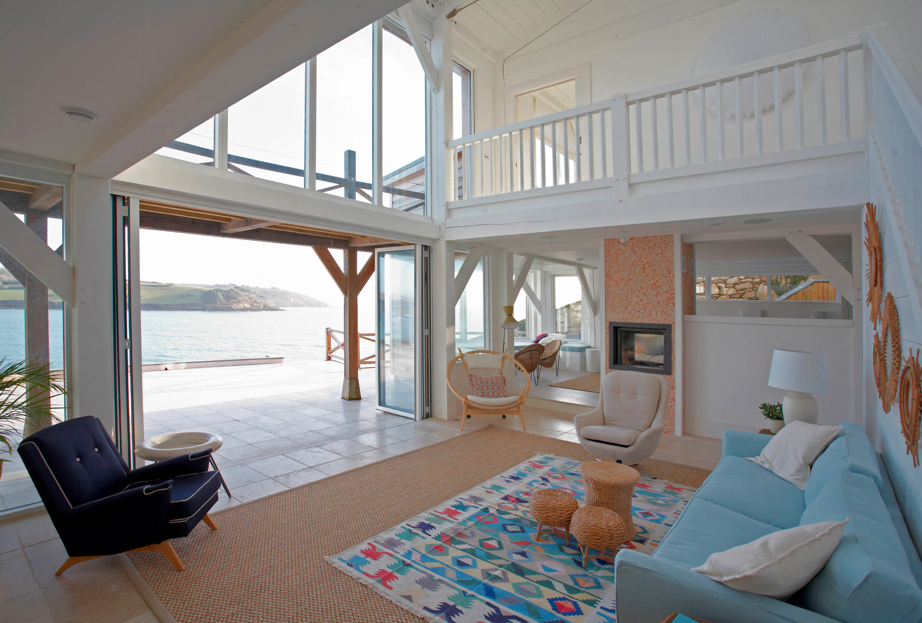 A double height living room in a coastal new built home in Cornwall with a white painted Douglas fir frame. A large sliding door is open with direct views onto the sea and opposite bay.