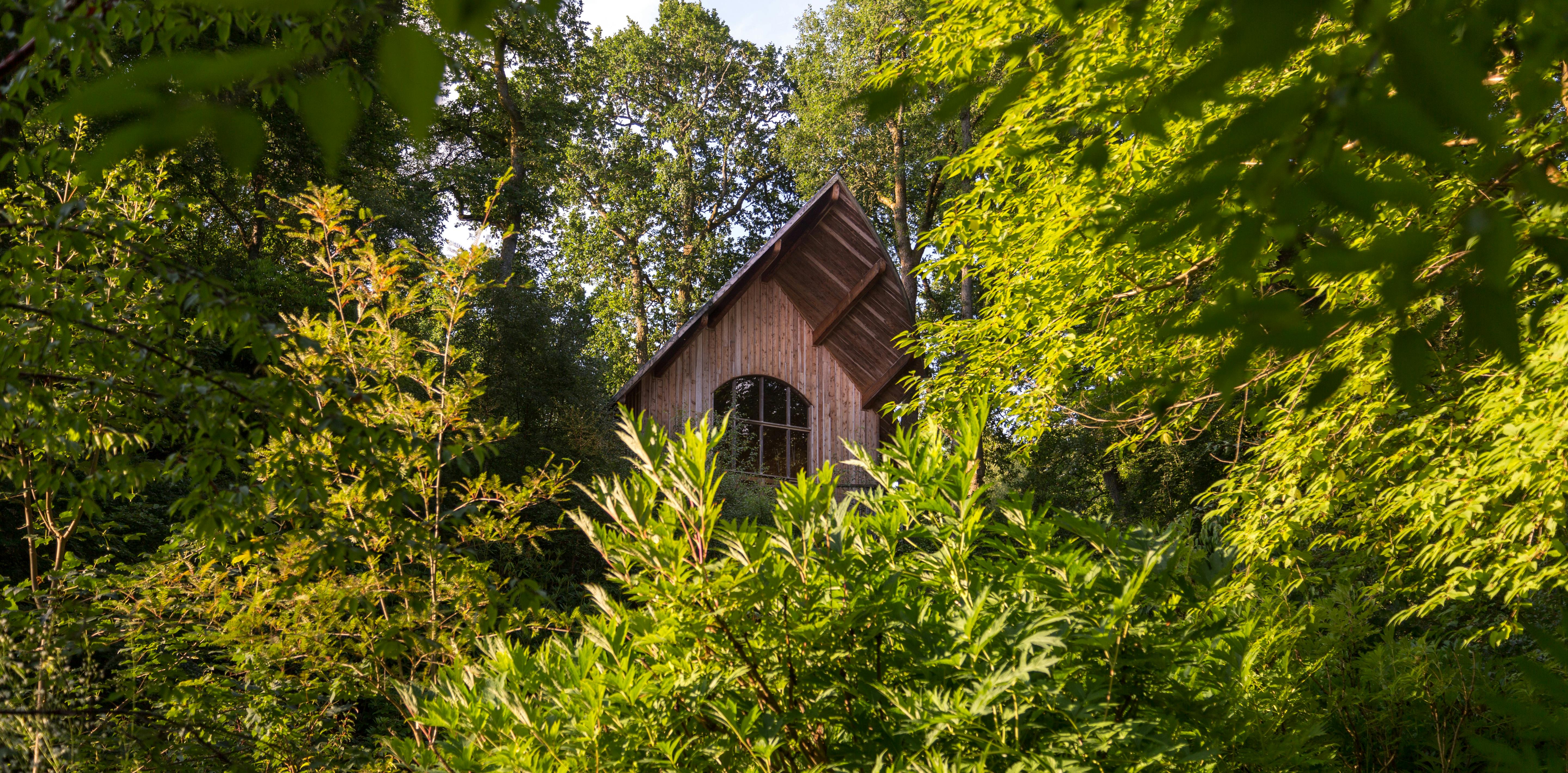 A wooden pavilion with an angled roof and a balcony can be seen between trees