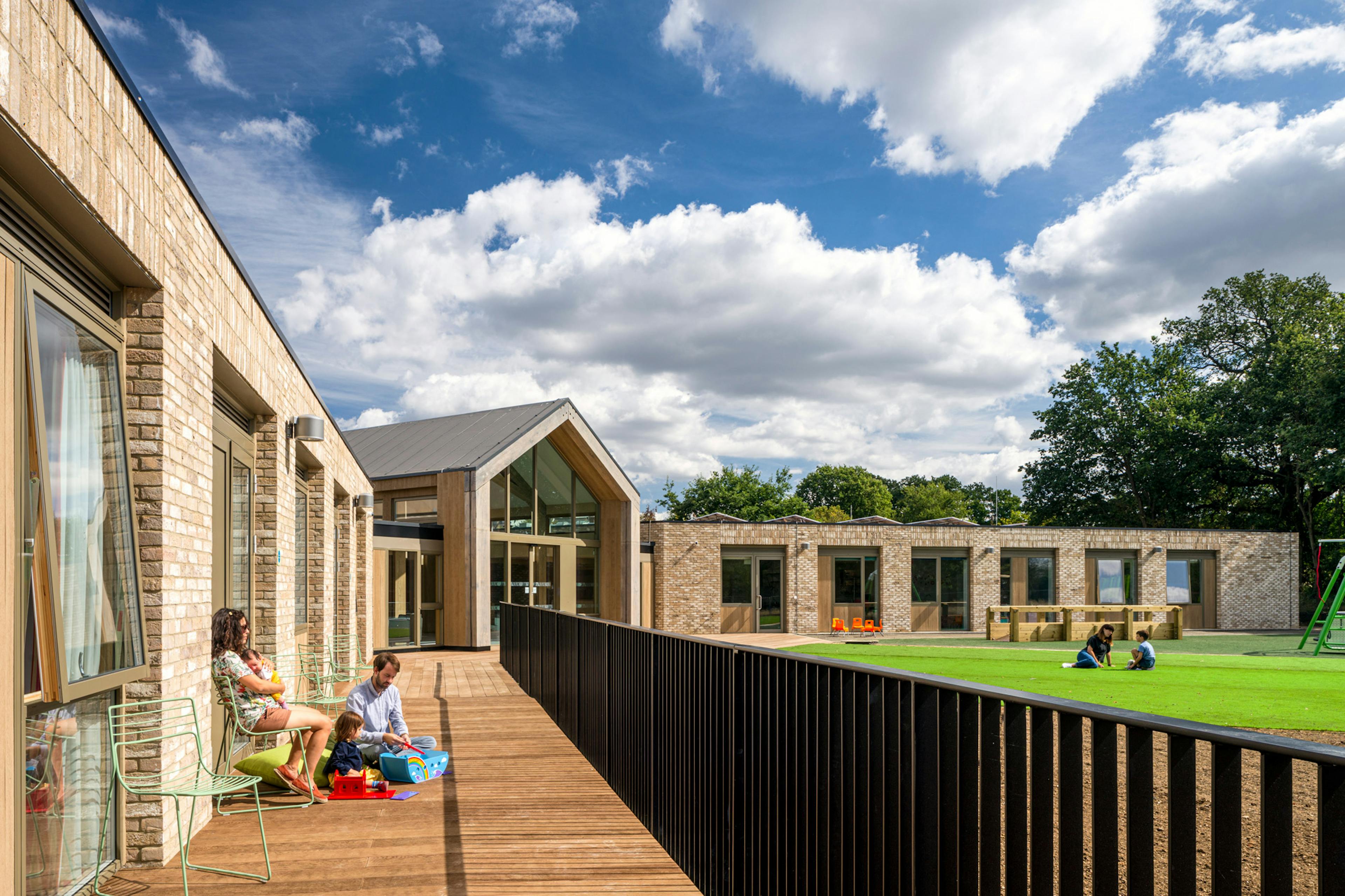 A family plays on a terrace outside a single storey brick children's hospital with a large oak framed atrium