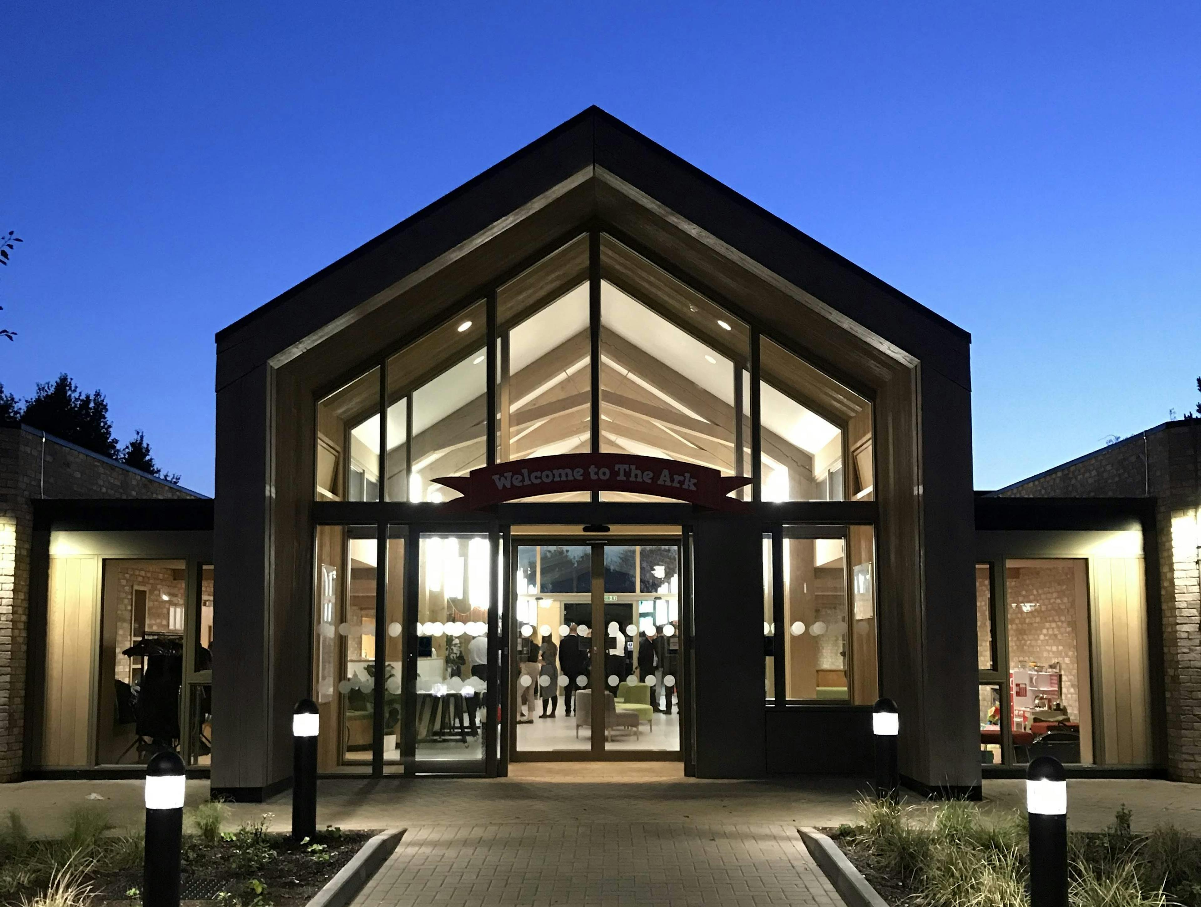At dusk lights shine from the windows of a single storey brick children's hospital with a large oak framed atrium