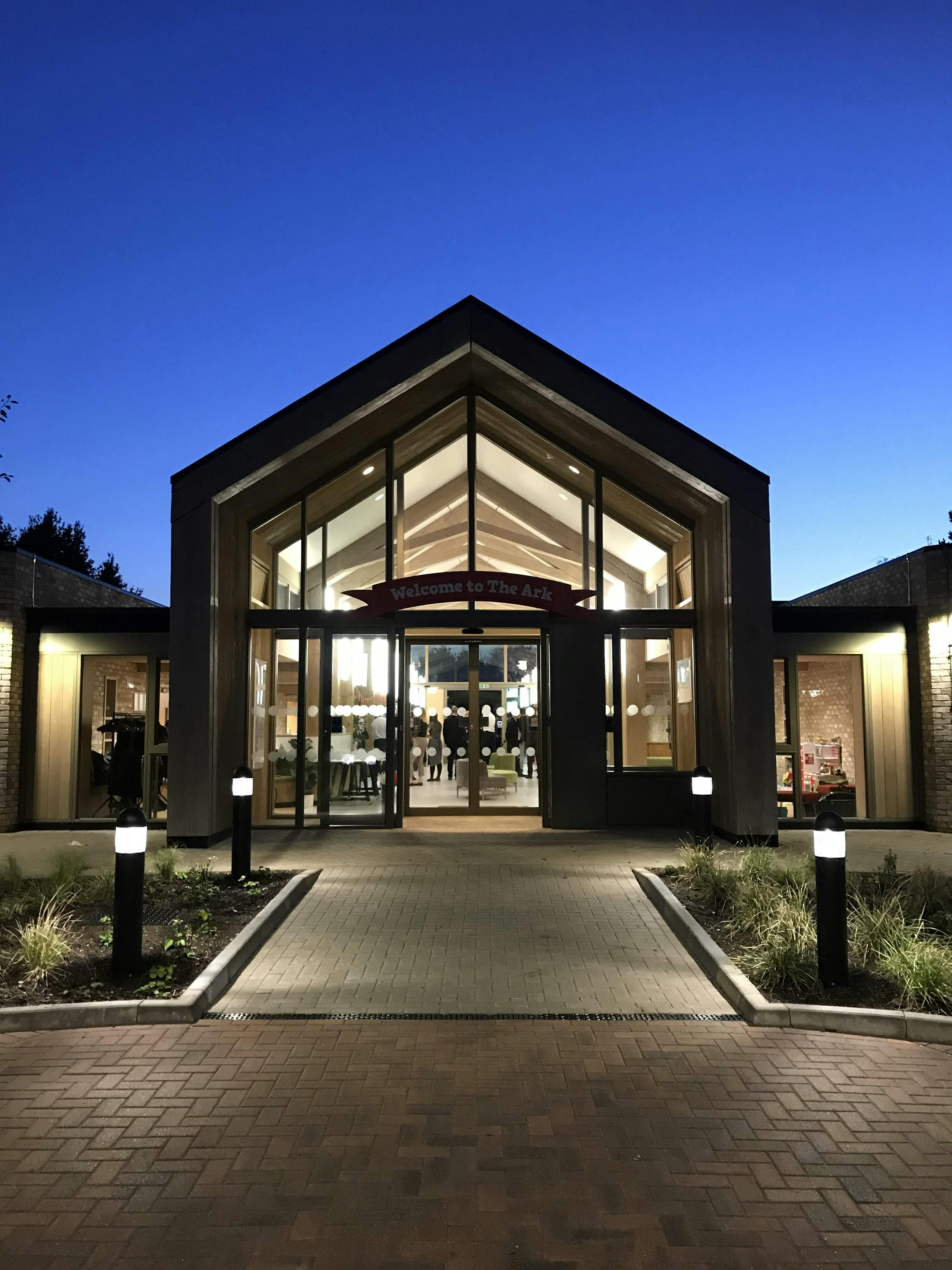 At dusk lights shine from the windows of a single storey brick children's hospital with a large oak framed atrium