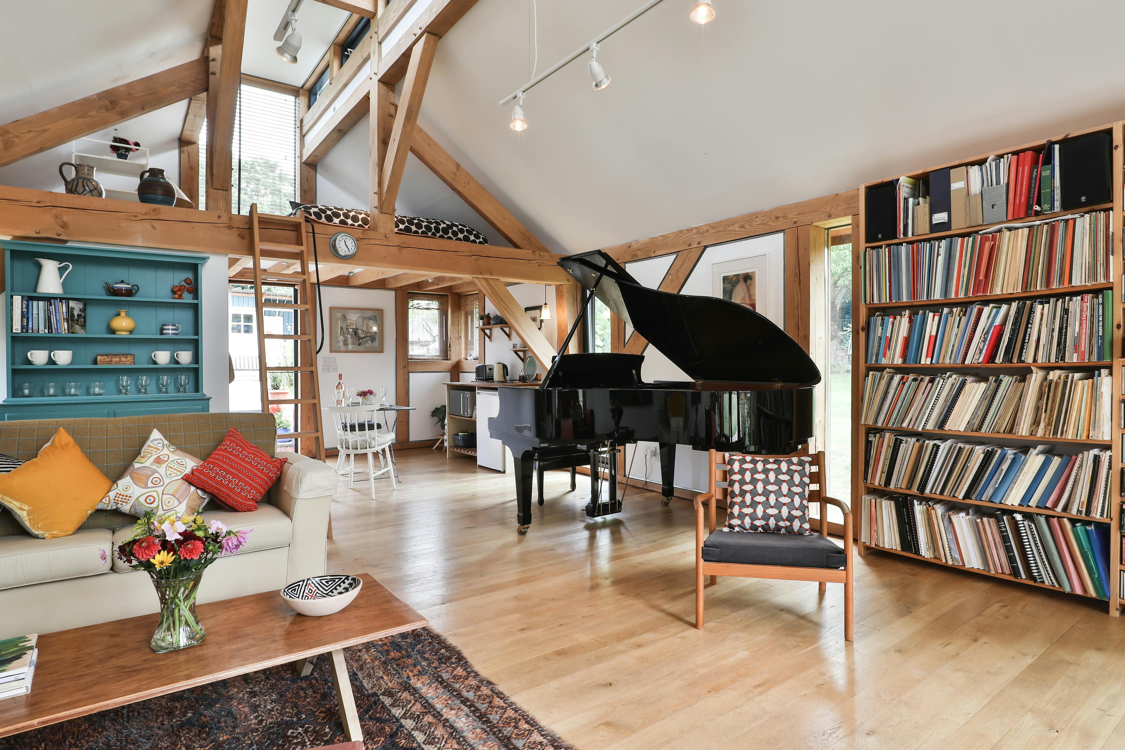 Sofas and a black grand piano in an open plan Douglas fir framed living room with a wooden ladder leading to a mezzanine level