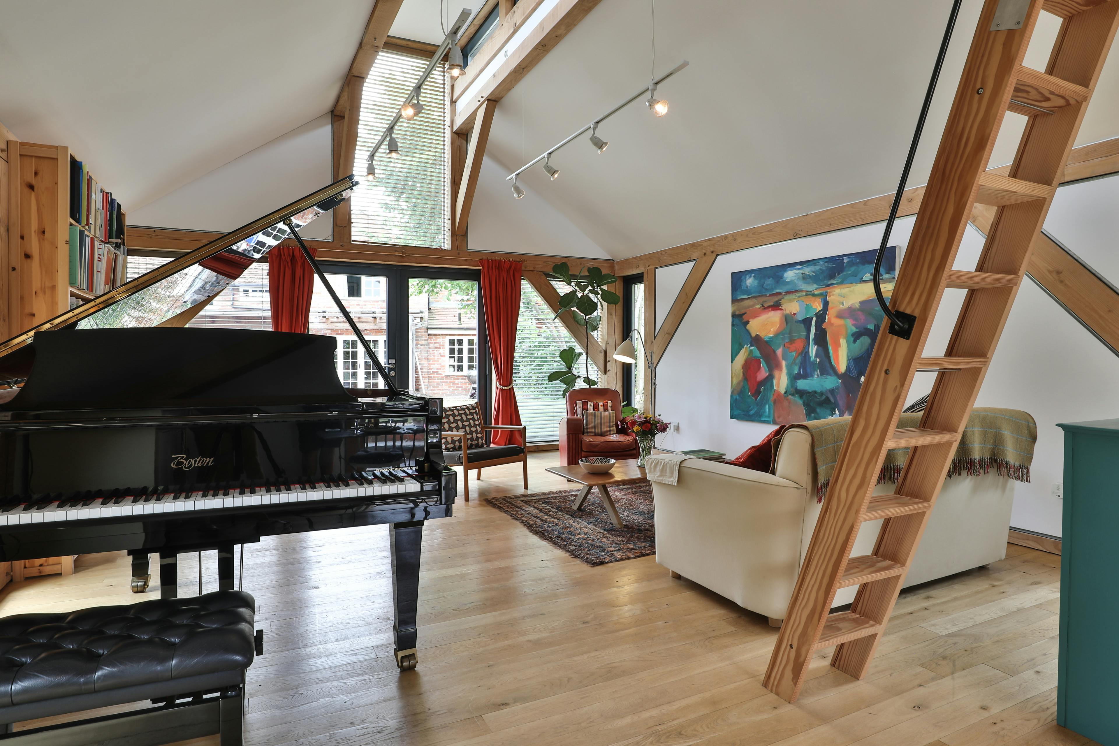 Sofas and a black grand piano in an open plan Douglas fir framed living room with a wooden ladder leading to a mezzanine space